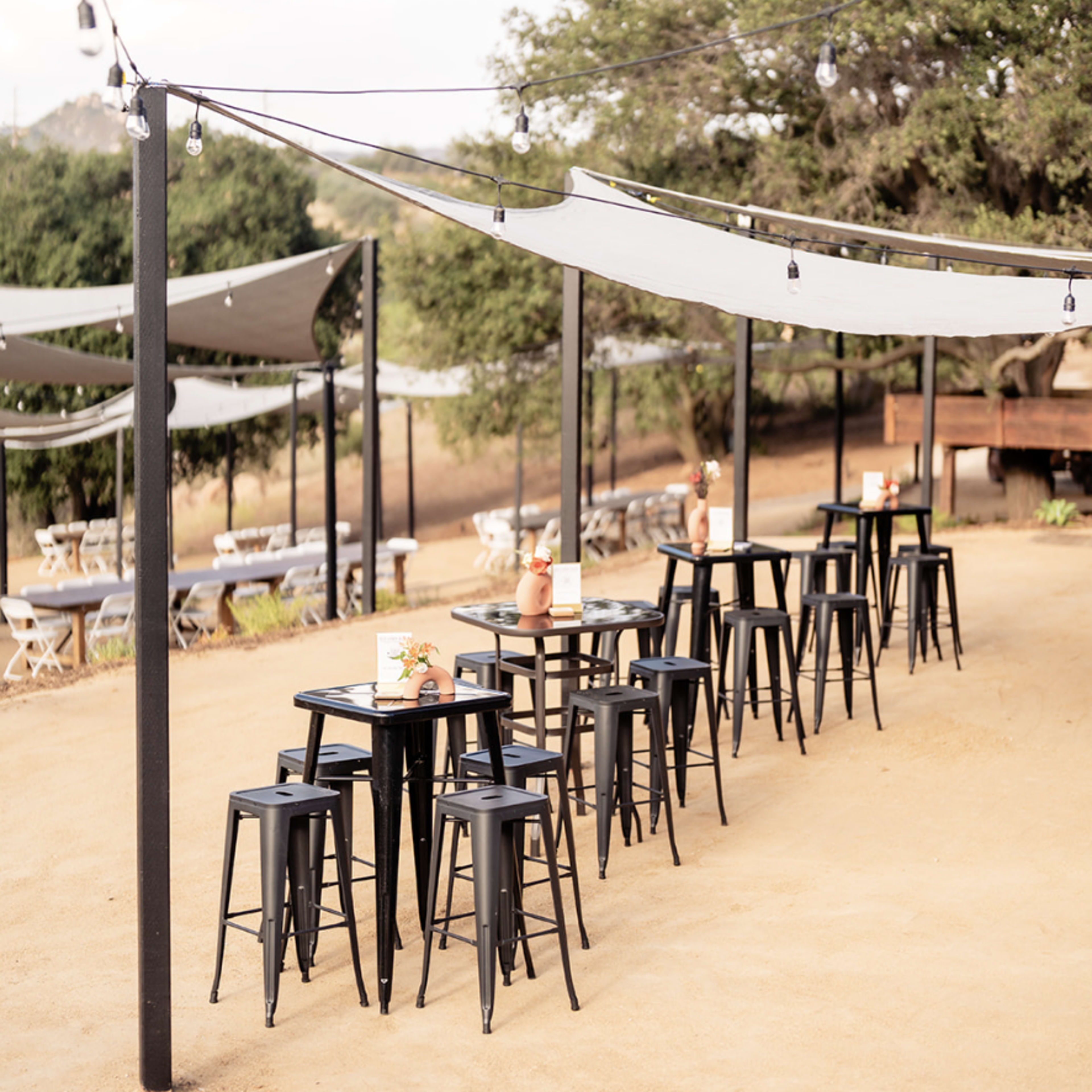 An outdoor dining area features rows of tall black stools and tables under shaded canopies, surrounded by trees.