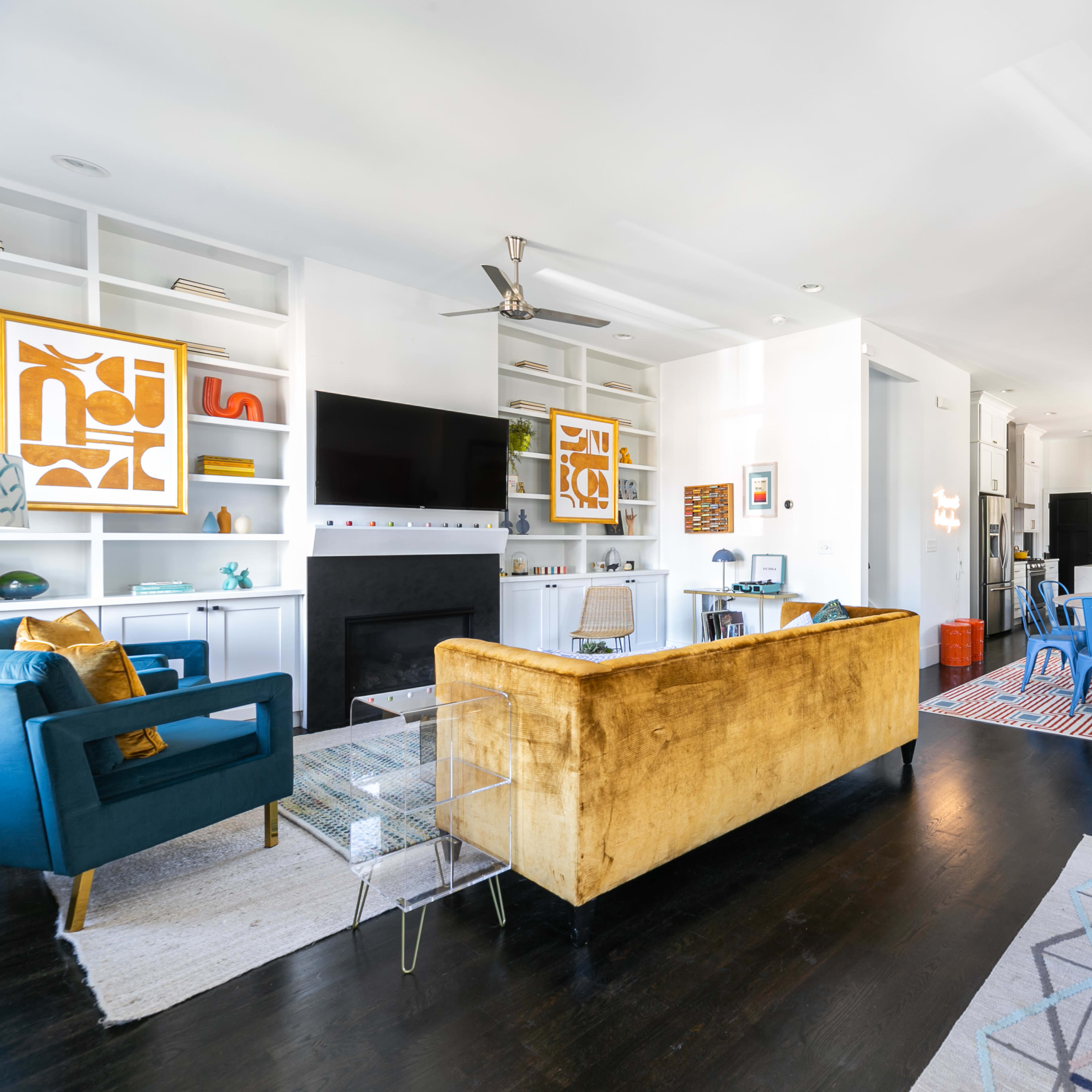 A bright, open living space features a mix of modern furniture, a television mounted on the wall, and a kitchen visible in the background.