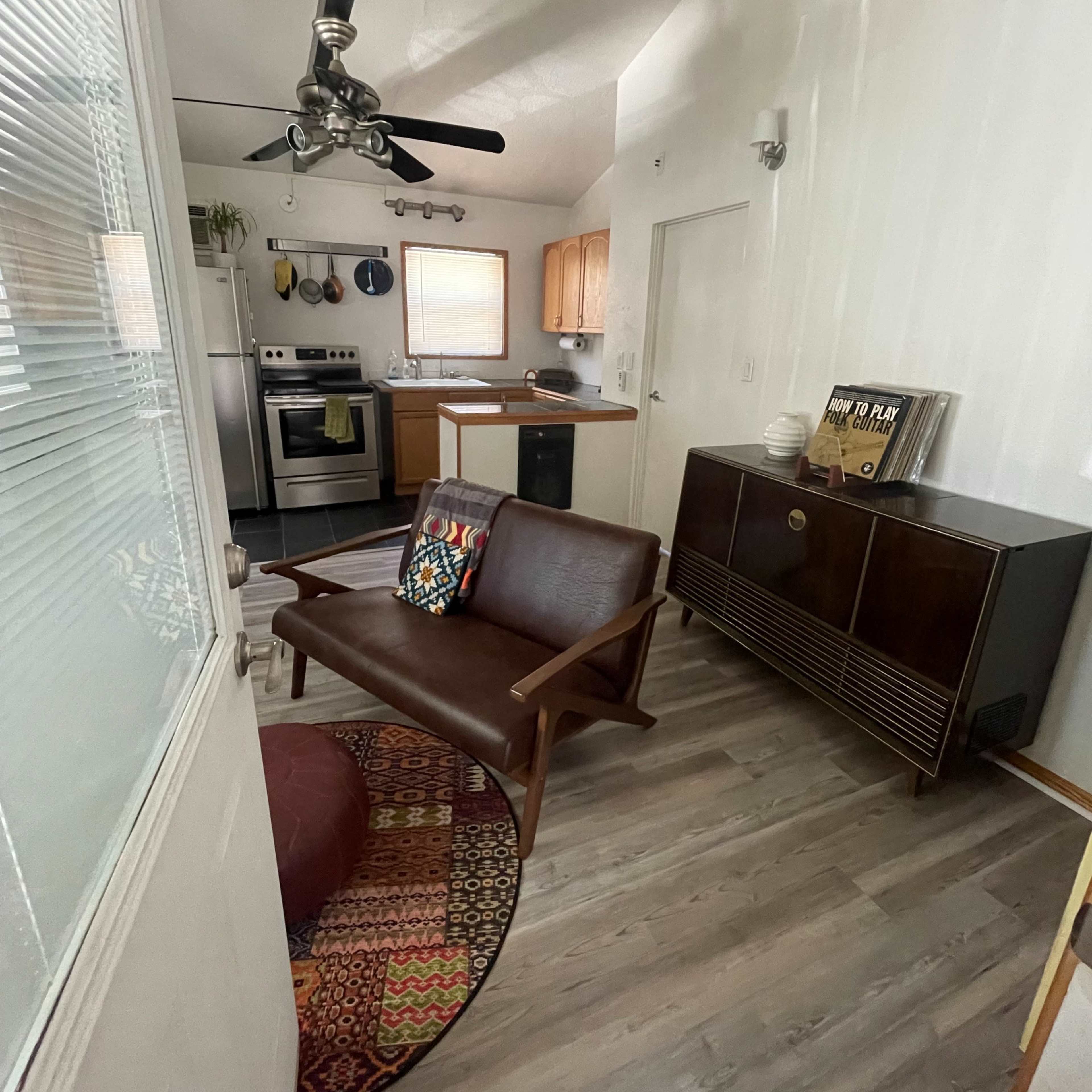 A small living area featuring a wooden couch, a round pouf, and a rug, with an open kitchen visible in the background.