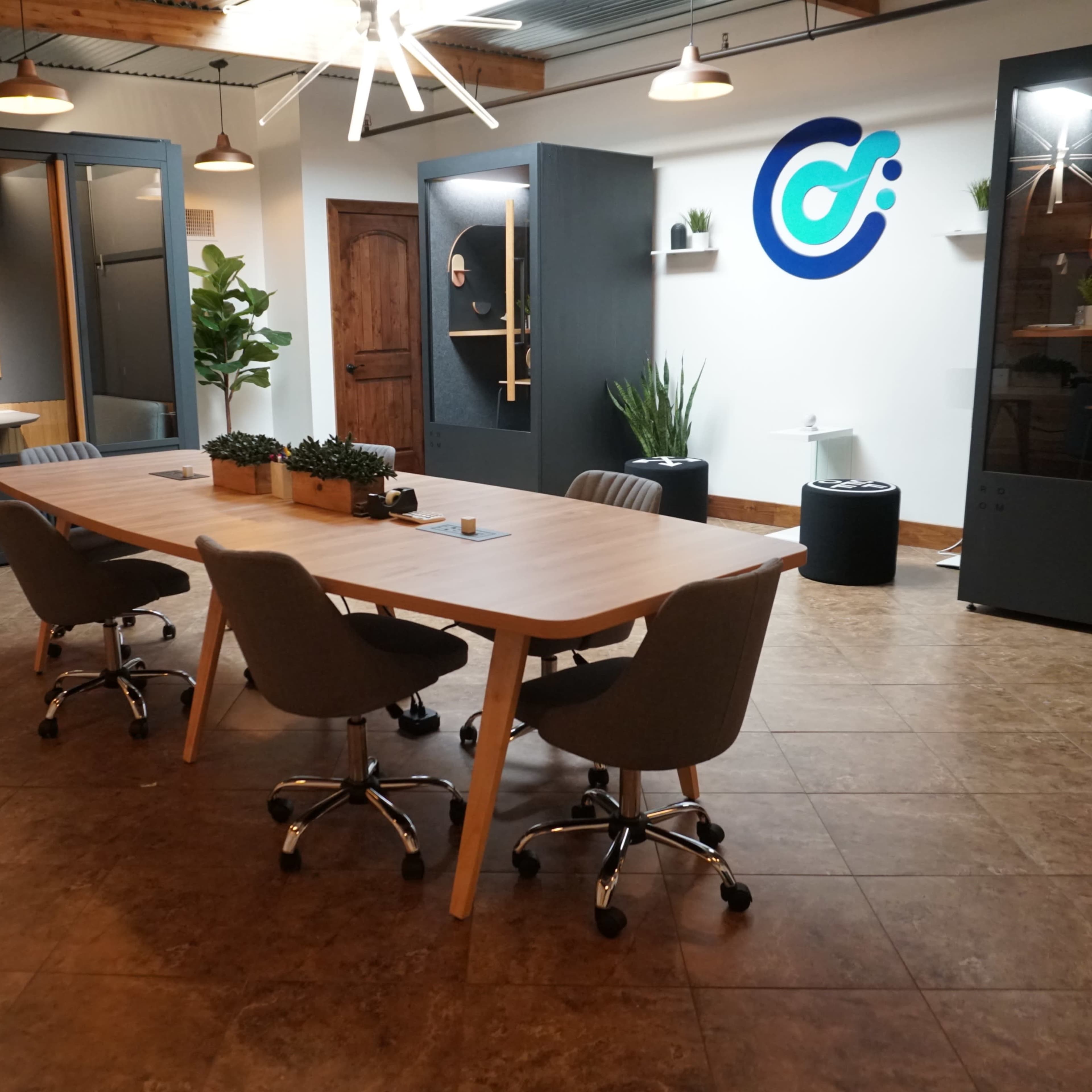 A modern office space featuring a long wooden table surrounded by ergonomic chairs, with glass pods and greenery in the background.
