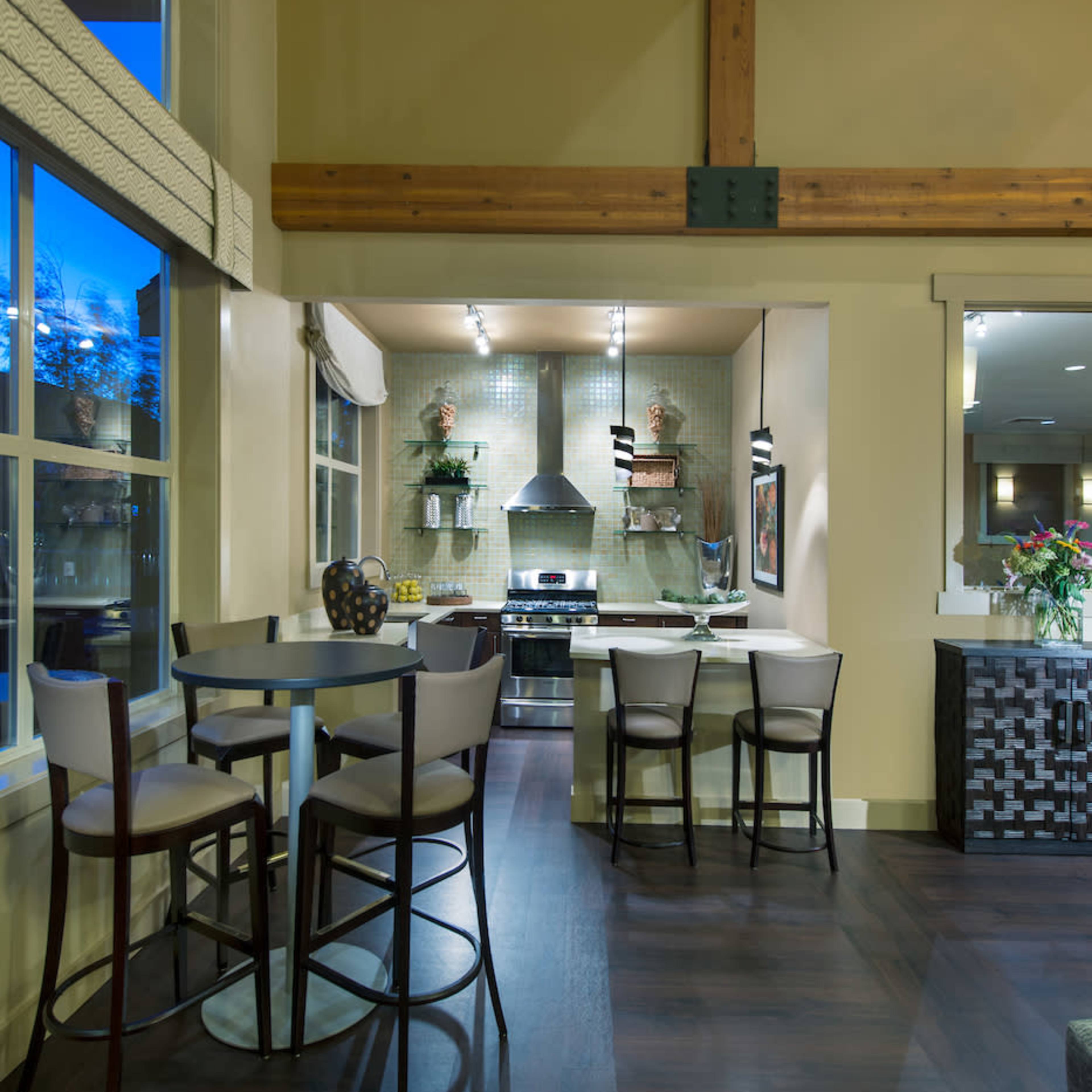 A modern kitchen with a dining area featuring tall bar stools, wooden beams, and a large window.