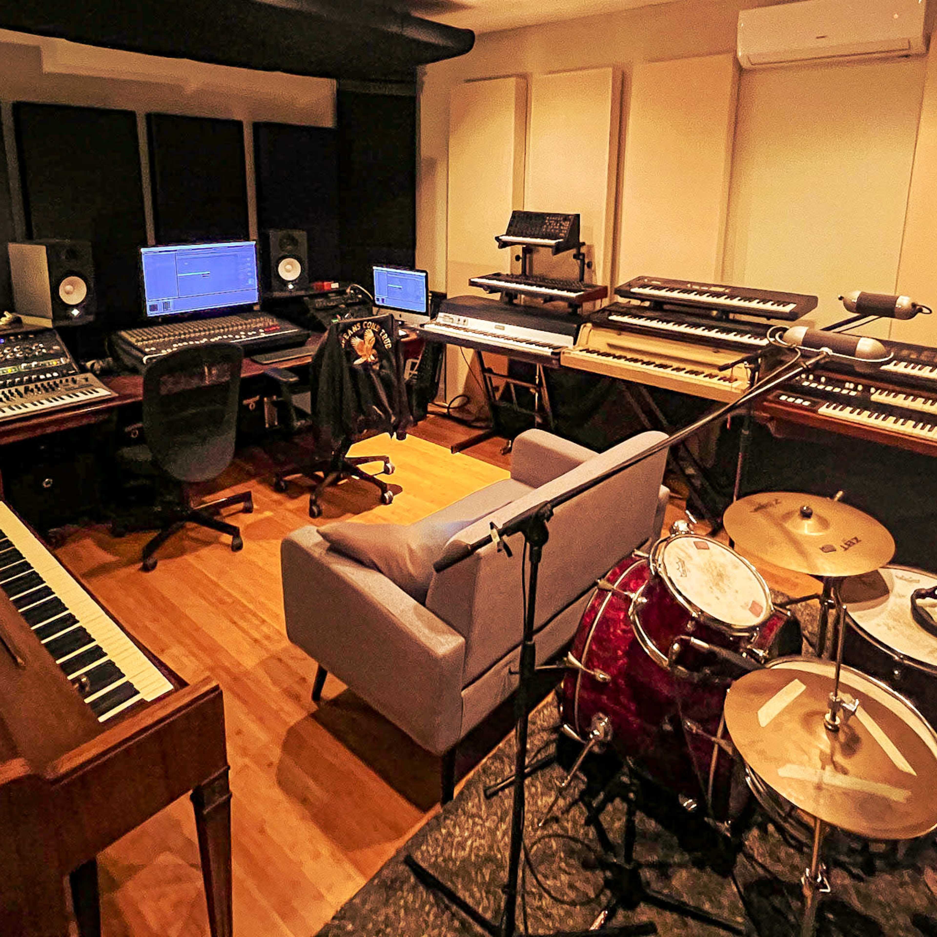 A music studio featuring a mixing console, multiple keyboards, a piano, a drum set, and seating arrangements.