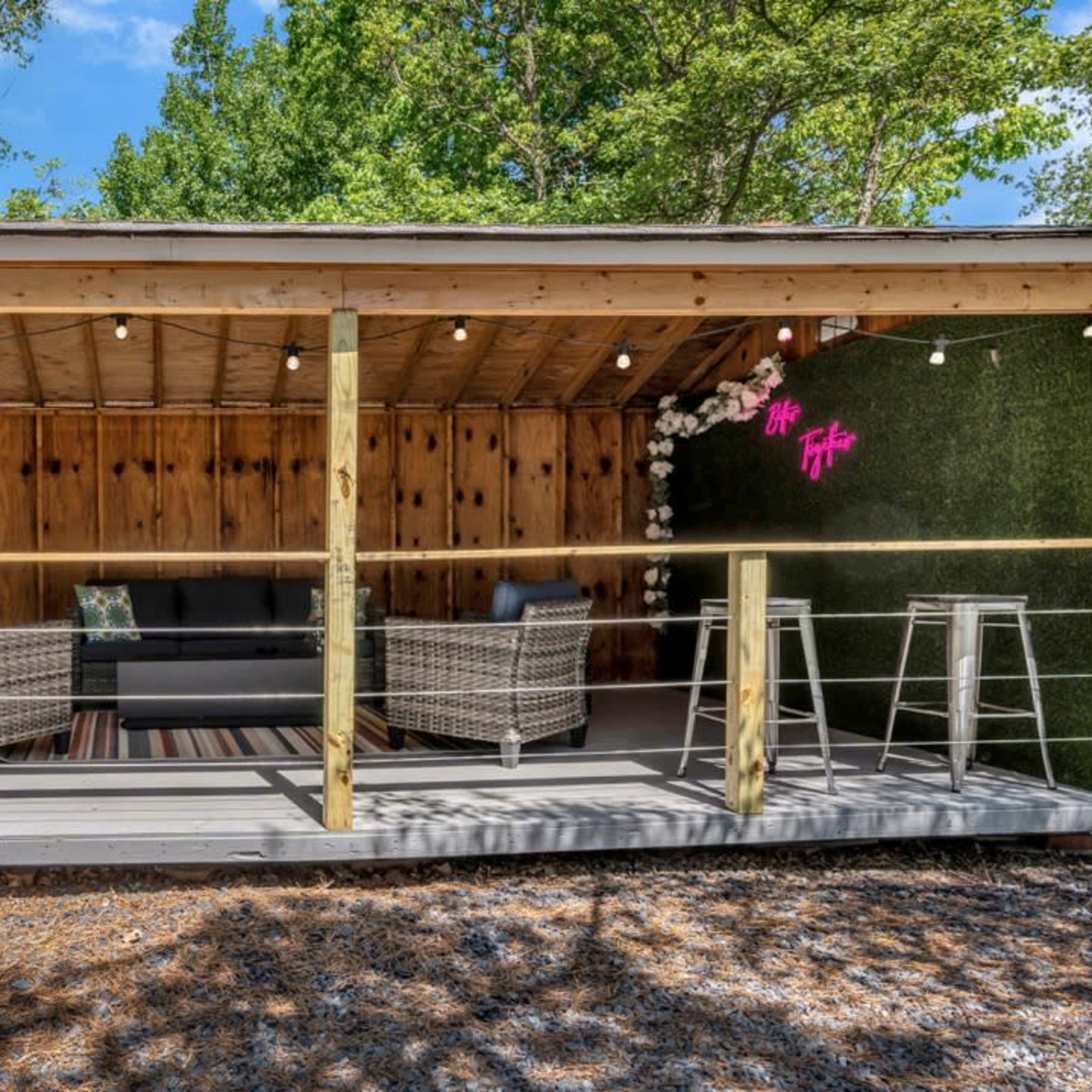 A modern outdoor lounge area with two wicker chairs, a sofa, and decorative plants under string lights, set within a wooden structure.
