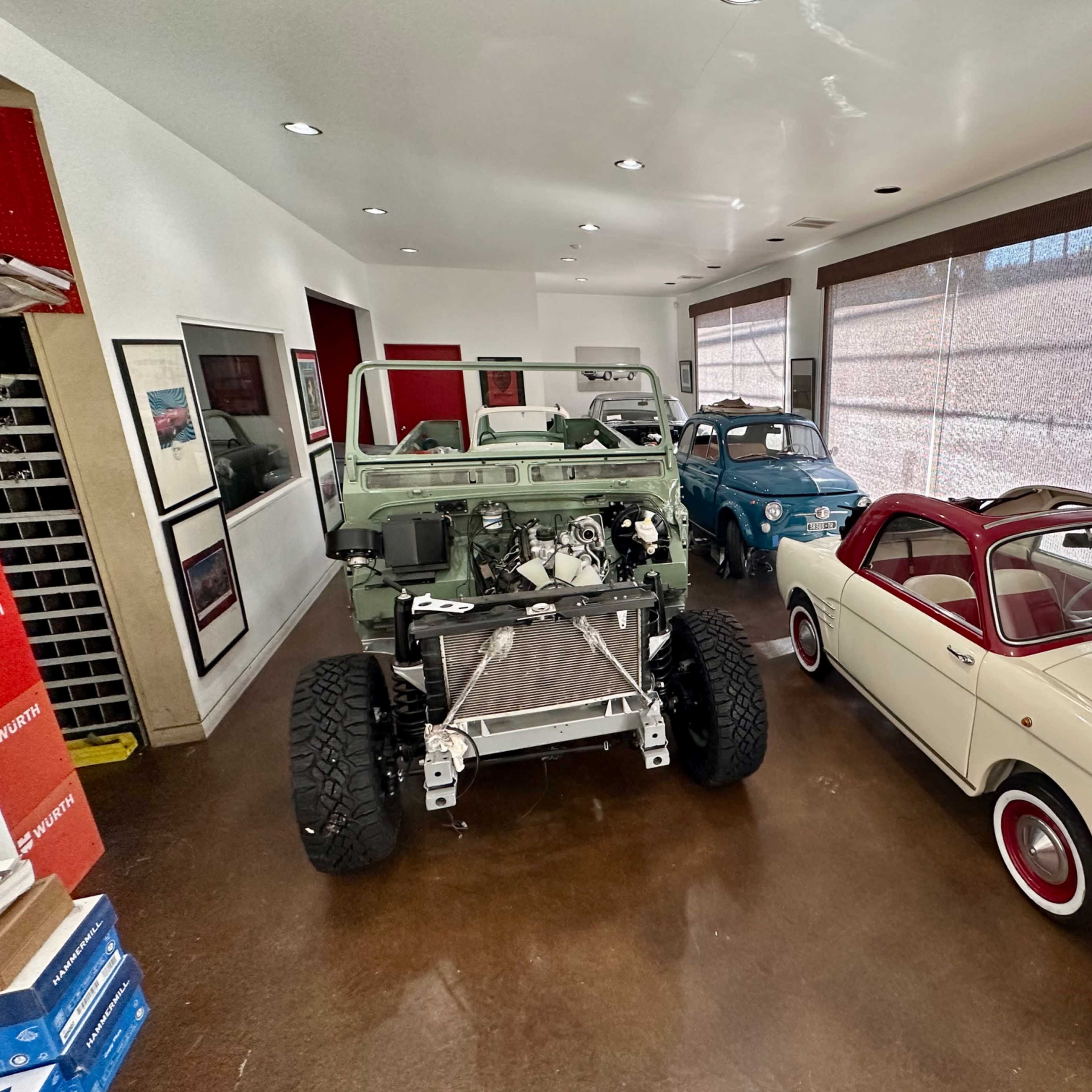 A narrow room with two distinct vintage cars on one side and an unfinished vehicle chassis on the other, with large windows letting in natural light.