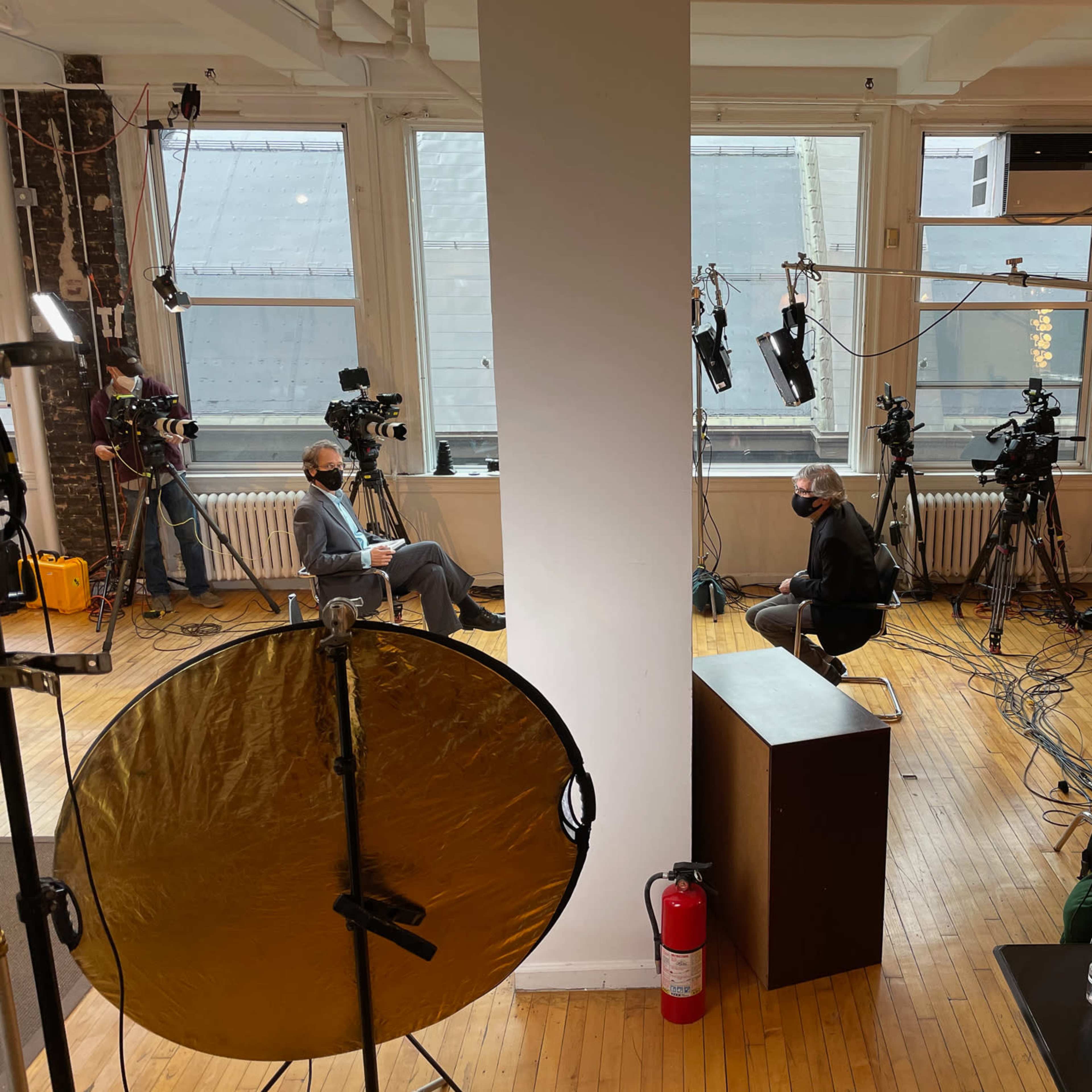 A photography studio is set up with multiple cameras and lighting equipment for a video shoot, featuring two individuals seated on opposite sides of a central dividing pillar.