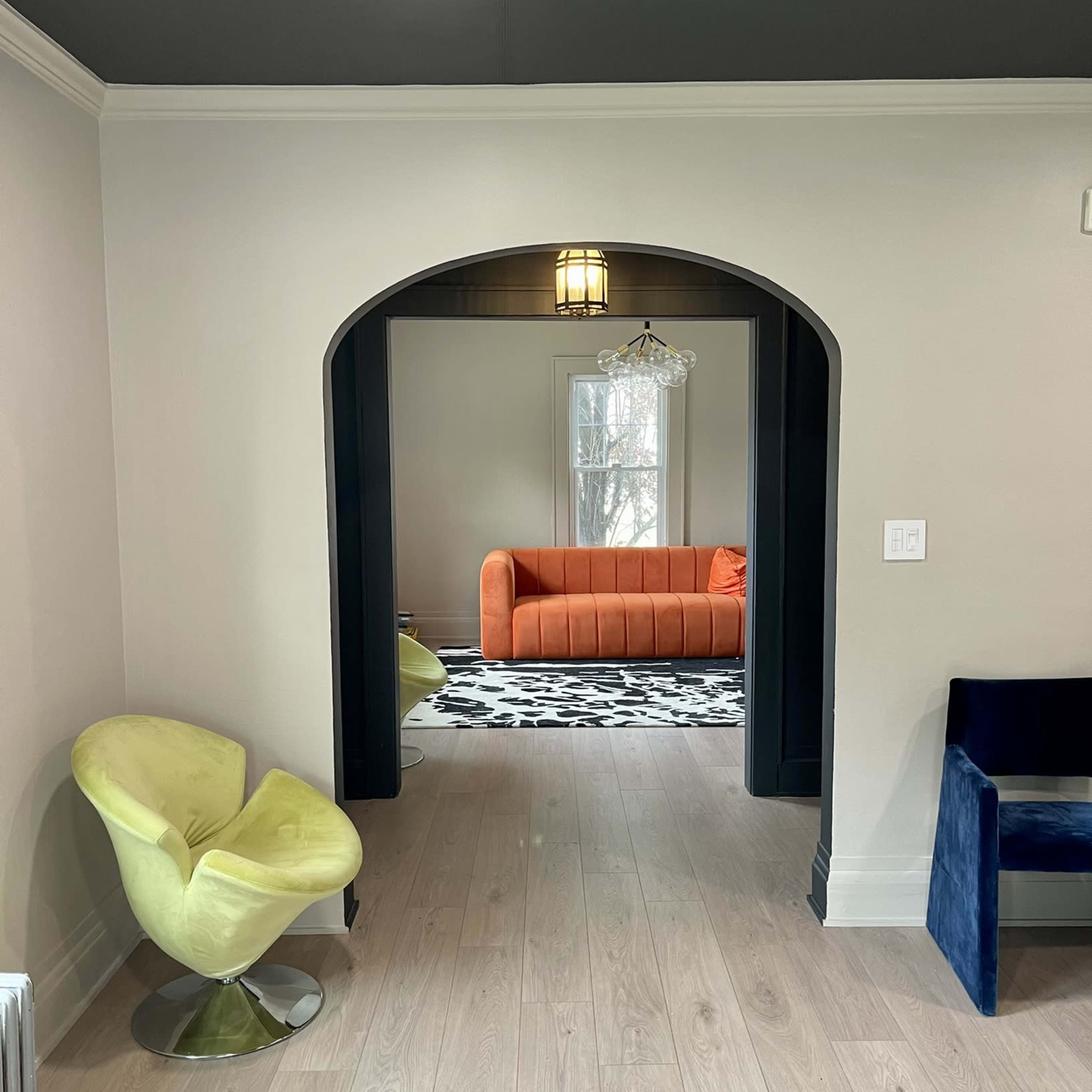 A brightly lit hallway with two modern chairs leads to a living room featuring an orange sofa and a geometric-patterned rug.