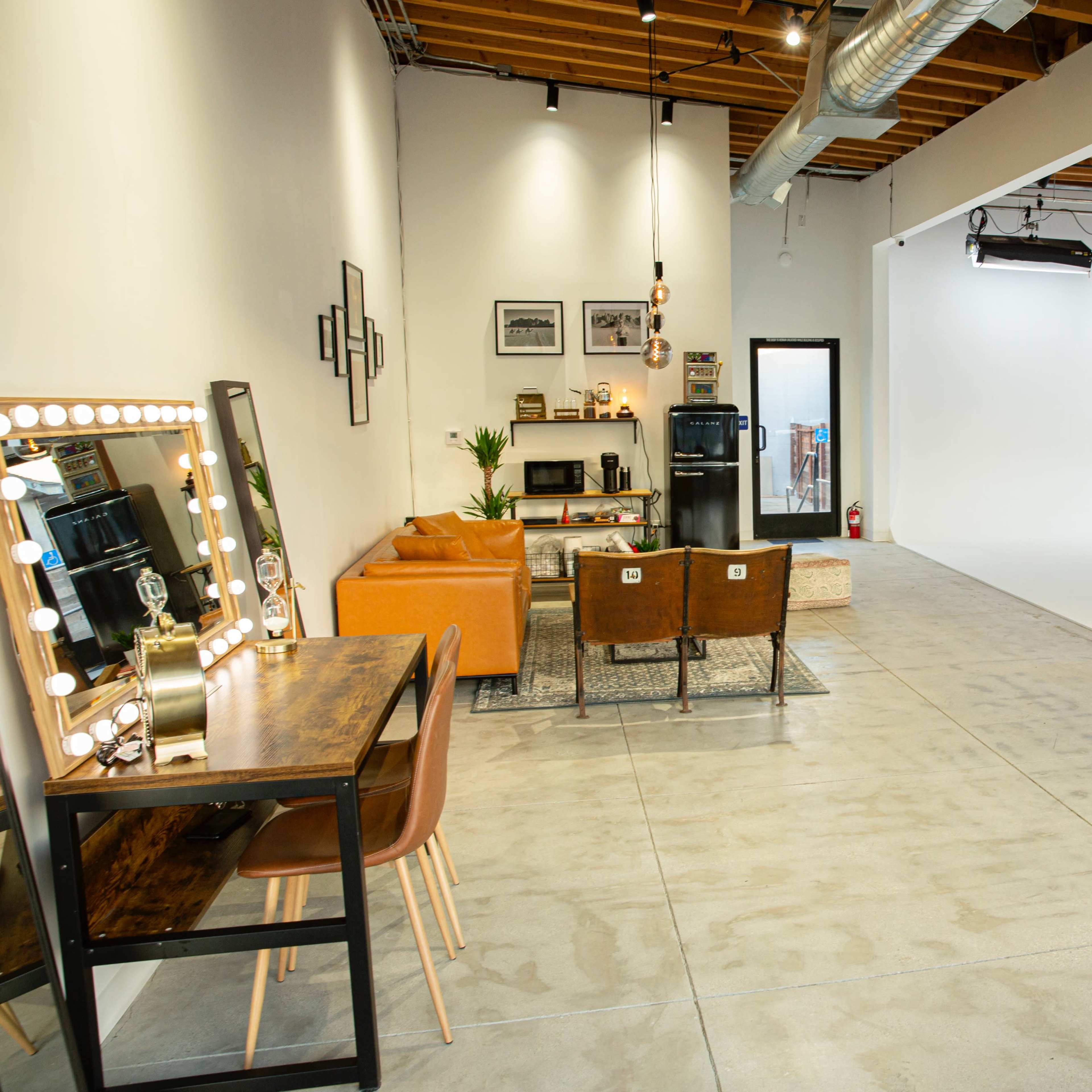 A spacious studio with a white backdrop, a seating area featuring a leather sofa and chairs, and a mirrored vanity table against one wall.