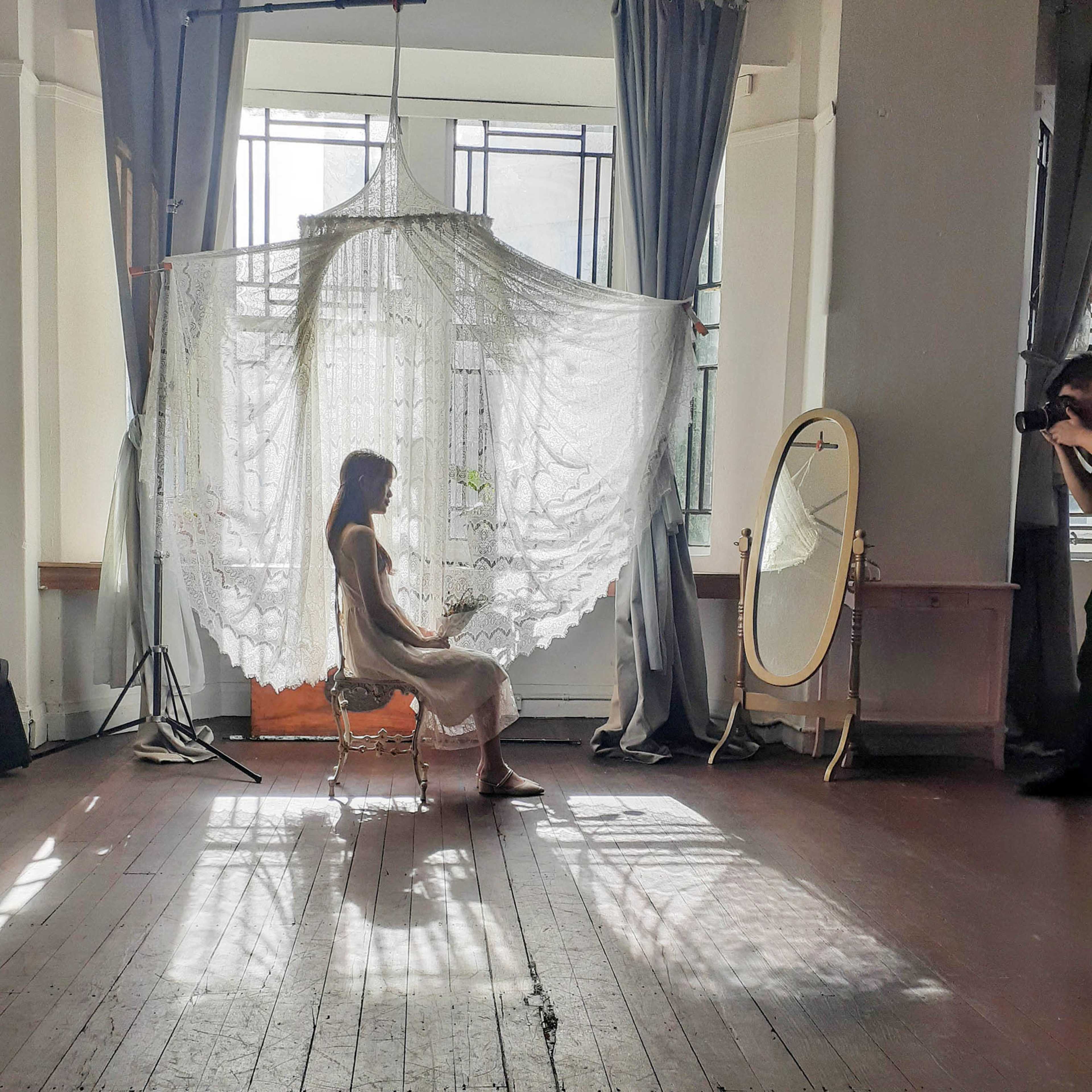 A woman sits on a chair in a well-lit room with a large lace curtain and an ornate mirror nearby, casting intricate shadows on the wooden floor.