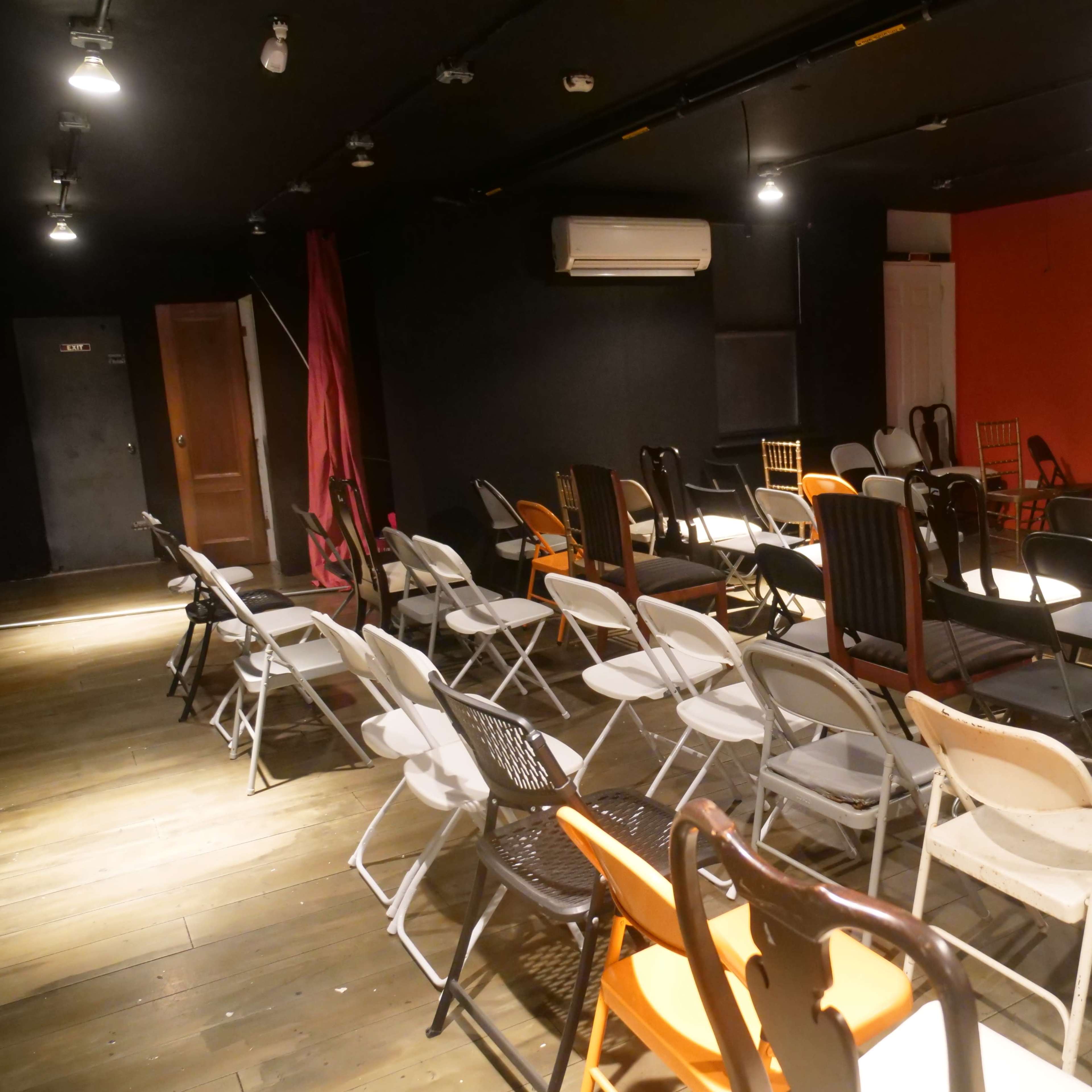 A room with various styles of chairs and tables arranged for a gathering, featuring dark walls and a bright orange accent wall.