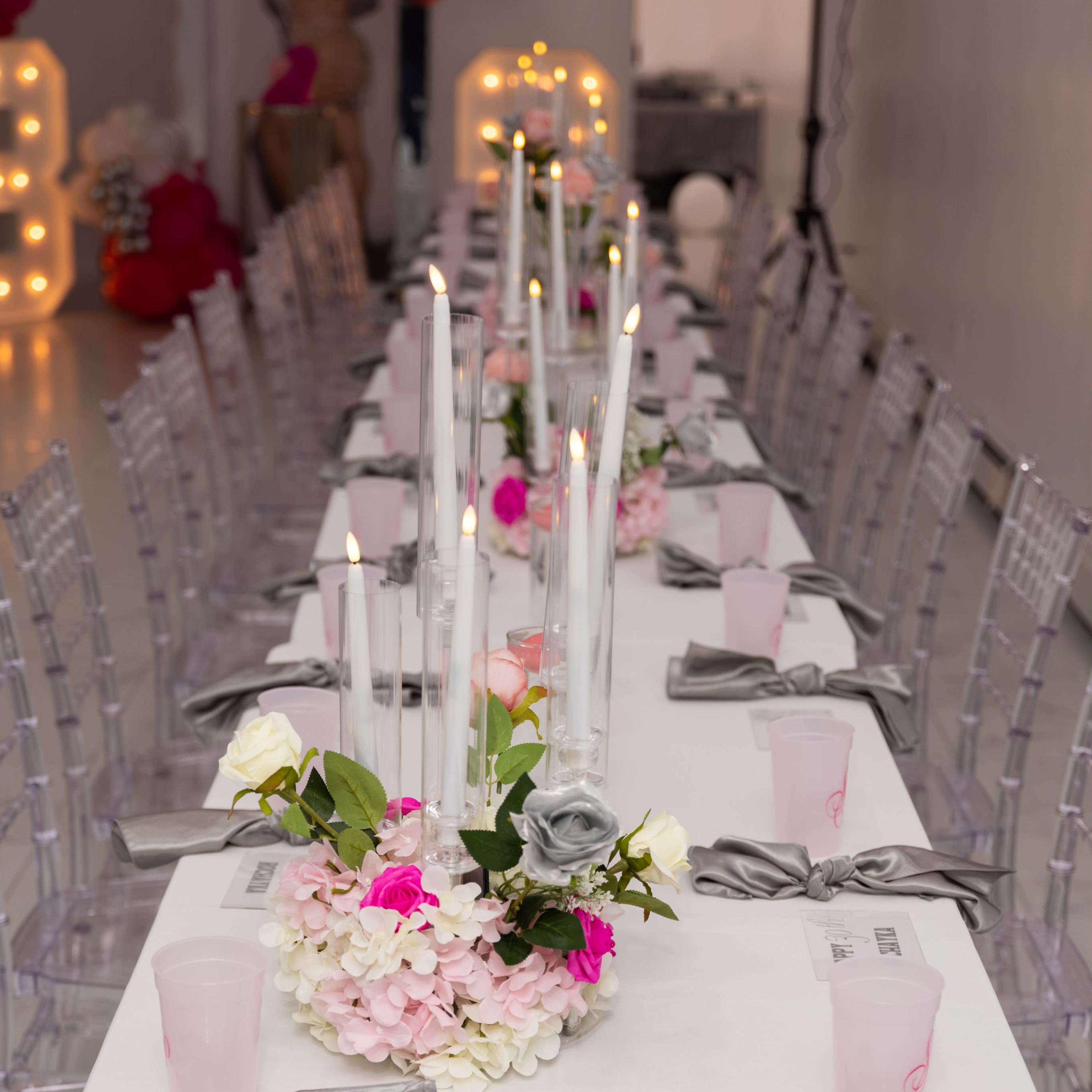 A long, elegantly set table features pink and white drinks cups, silver bows on white napkins, and tall candles surrounded by floral arrangements.