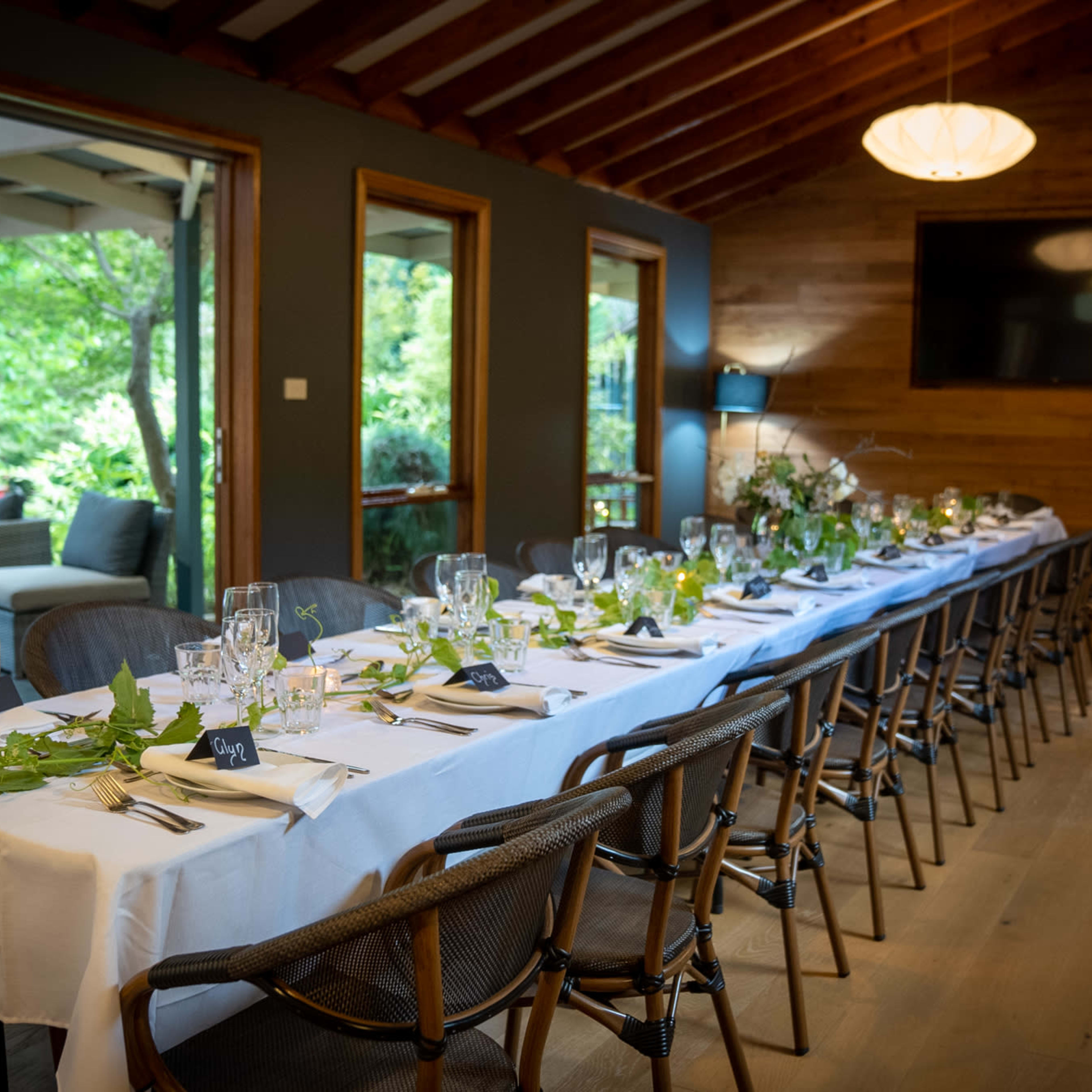 A long dining table is elegantly set with tableware and greenery in a rustic dining room overlooking a lush outdoor area.