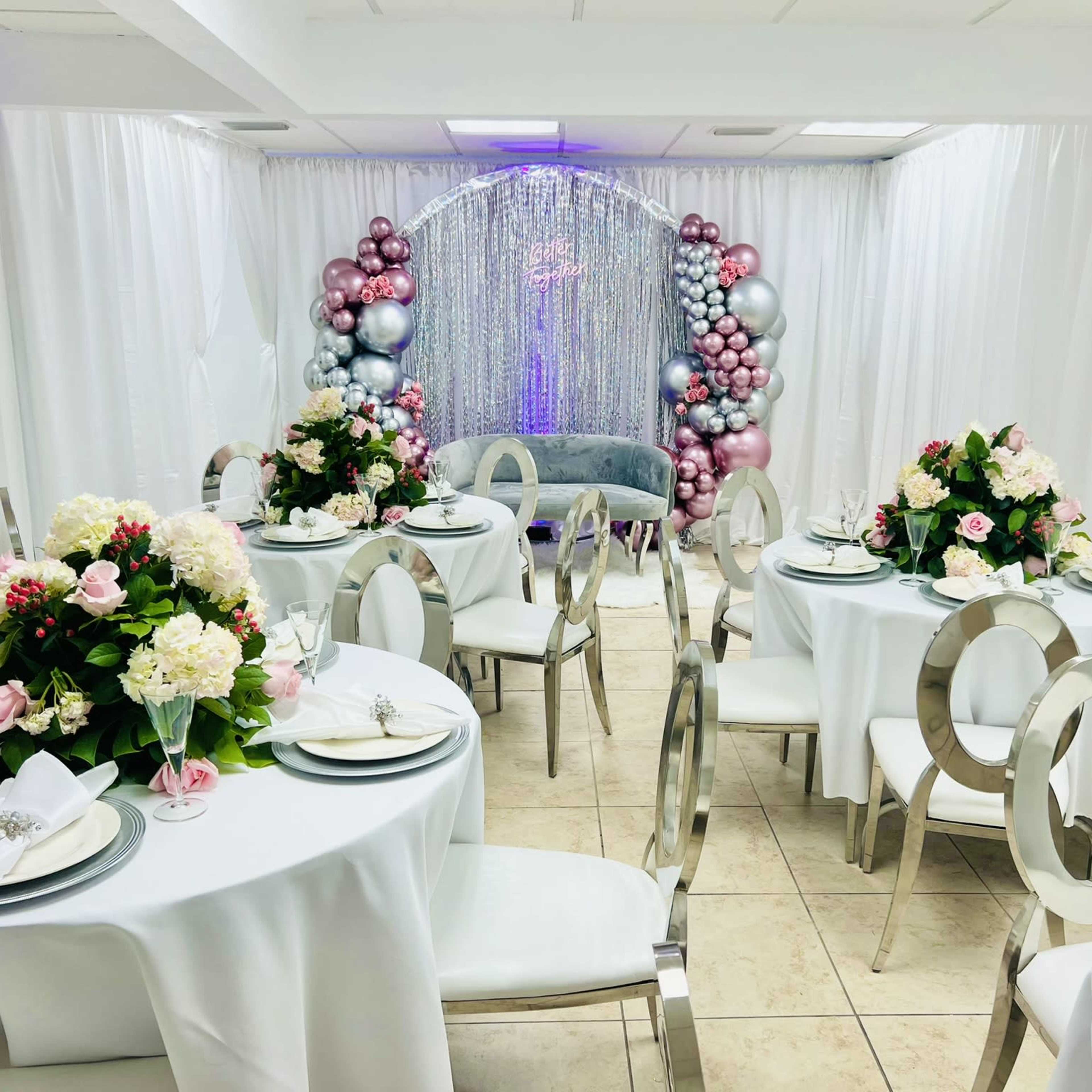 A decorated indoor venue with round tables, floral centerpieces, and a backdrop of balloons and shimmering fabric.