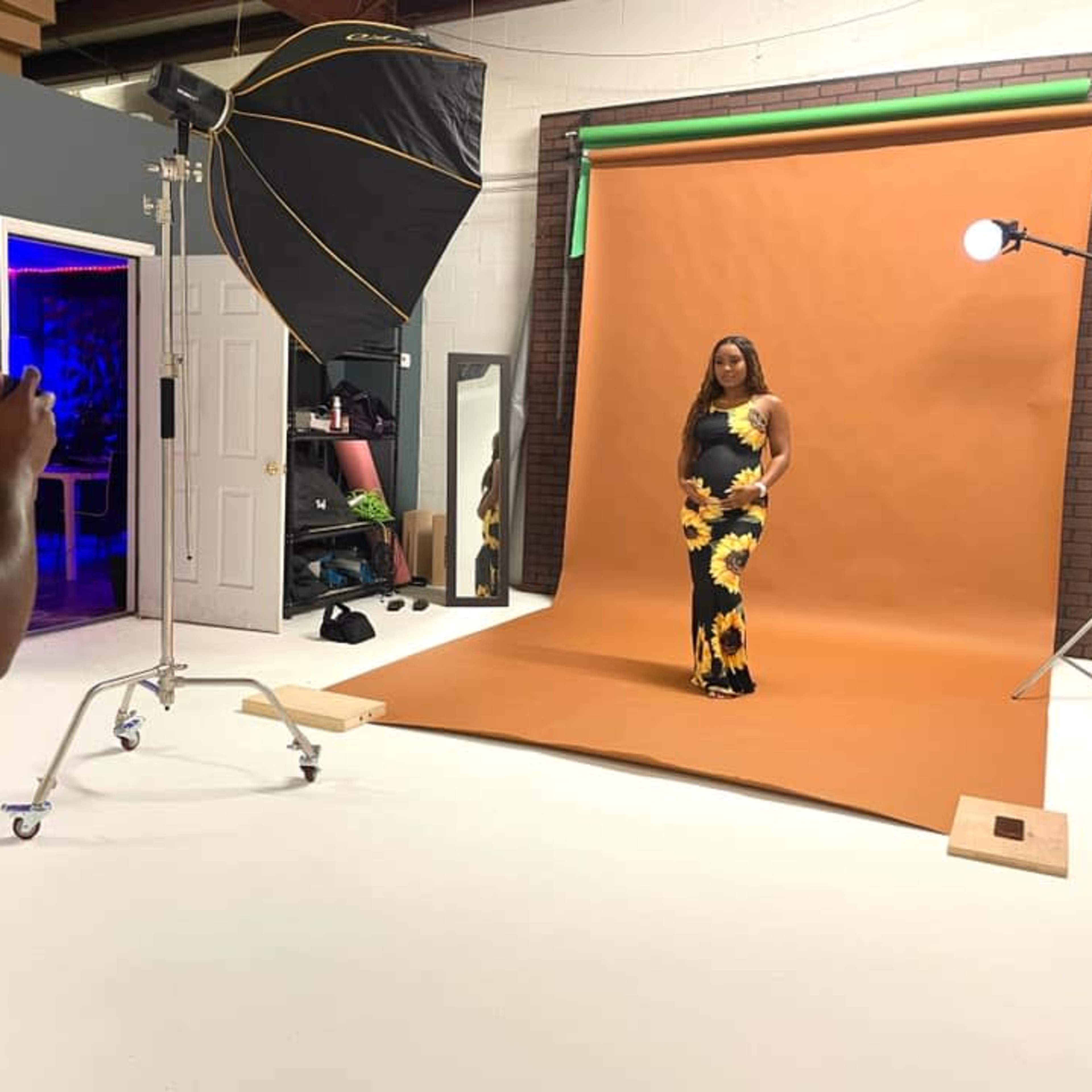 A photographer takes pictures of a model in a sunflower-patterned dress against a brown backdrop in a studio.
