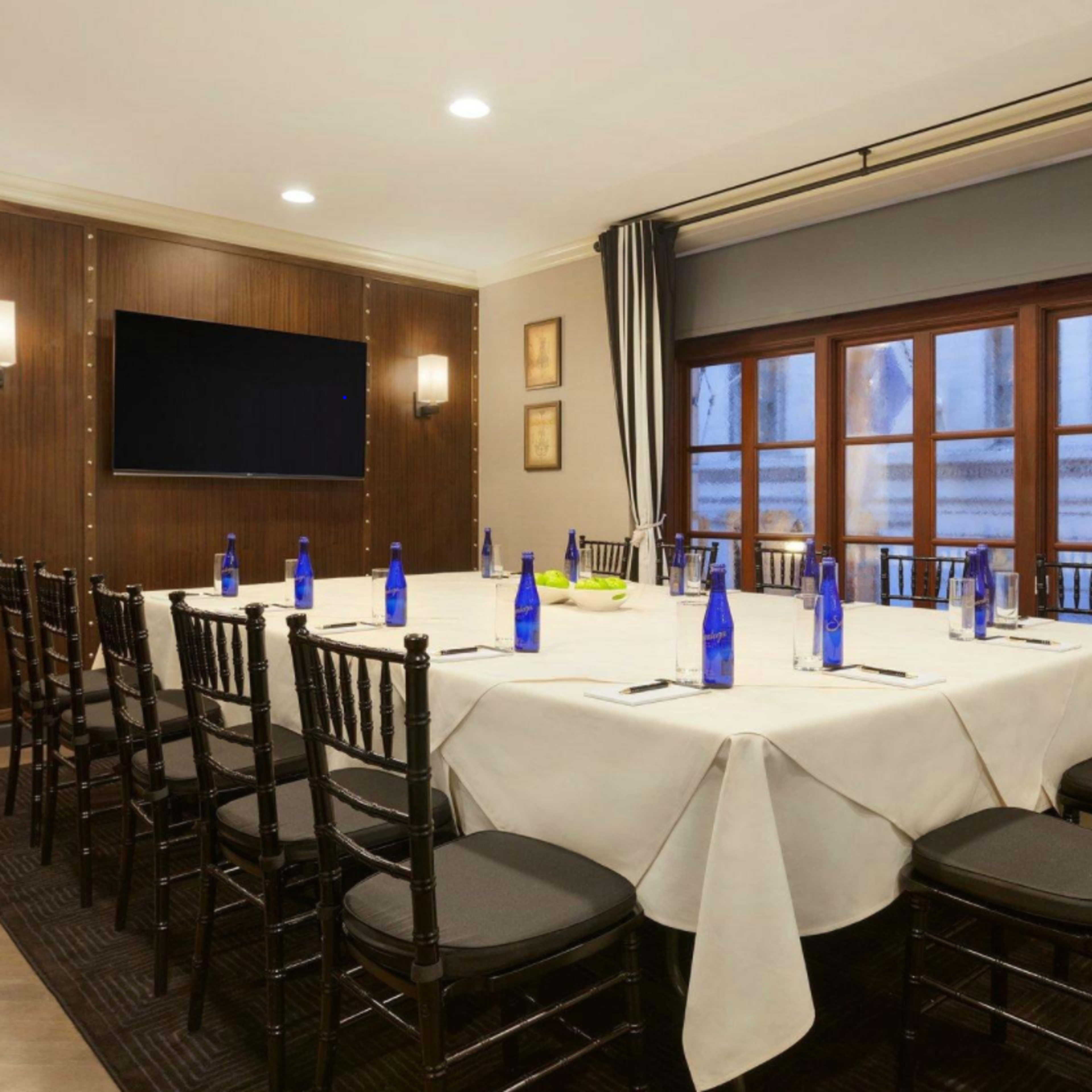 A neatly arranged conference room features a round table surrounded by black chairs, with blue water bottles placed at each seat and large windows providing light.