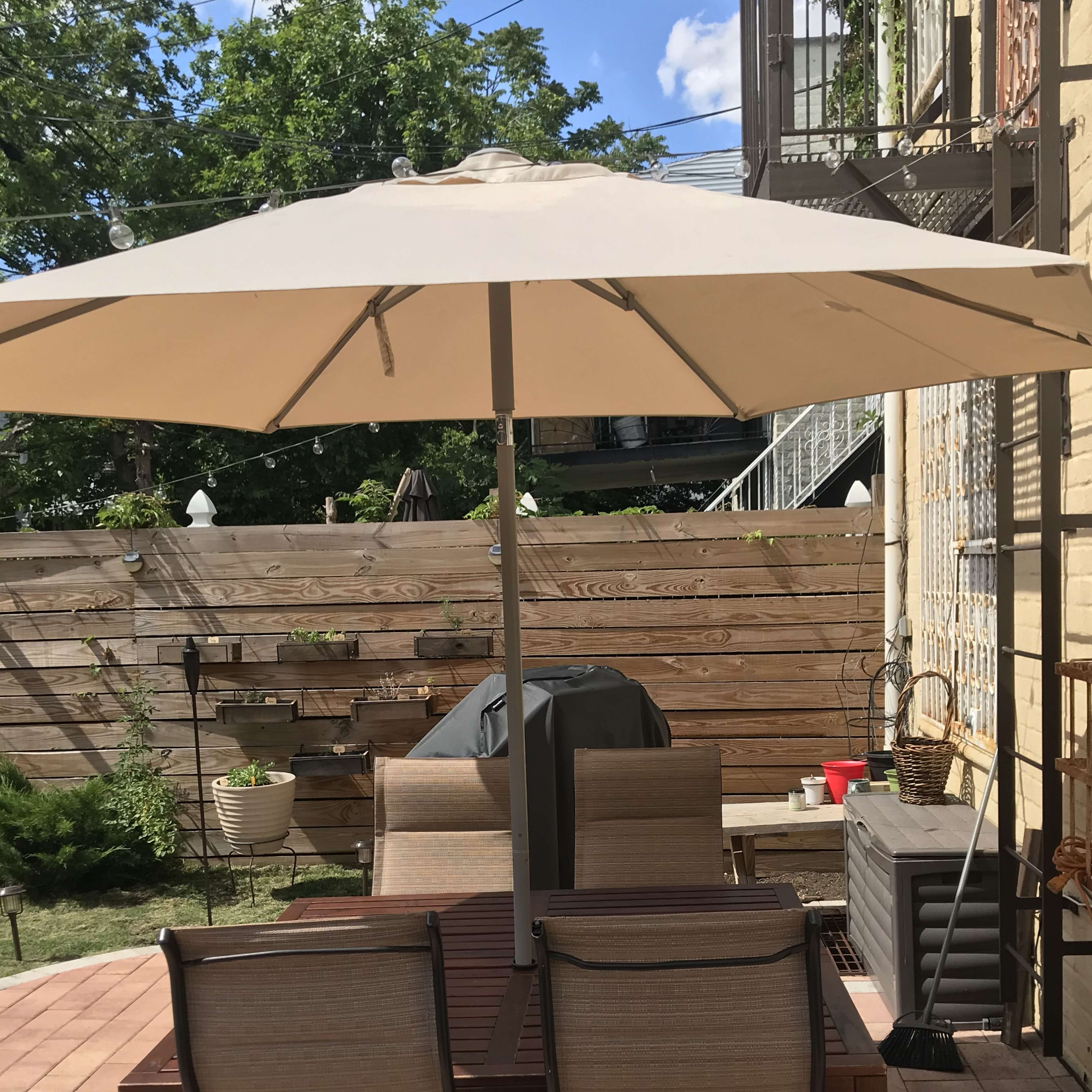 A patio area features a table with several chairs under an umbrella, surrounded by a wooden fence and potted plants.