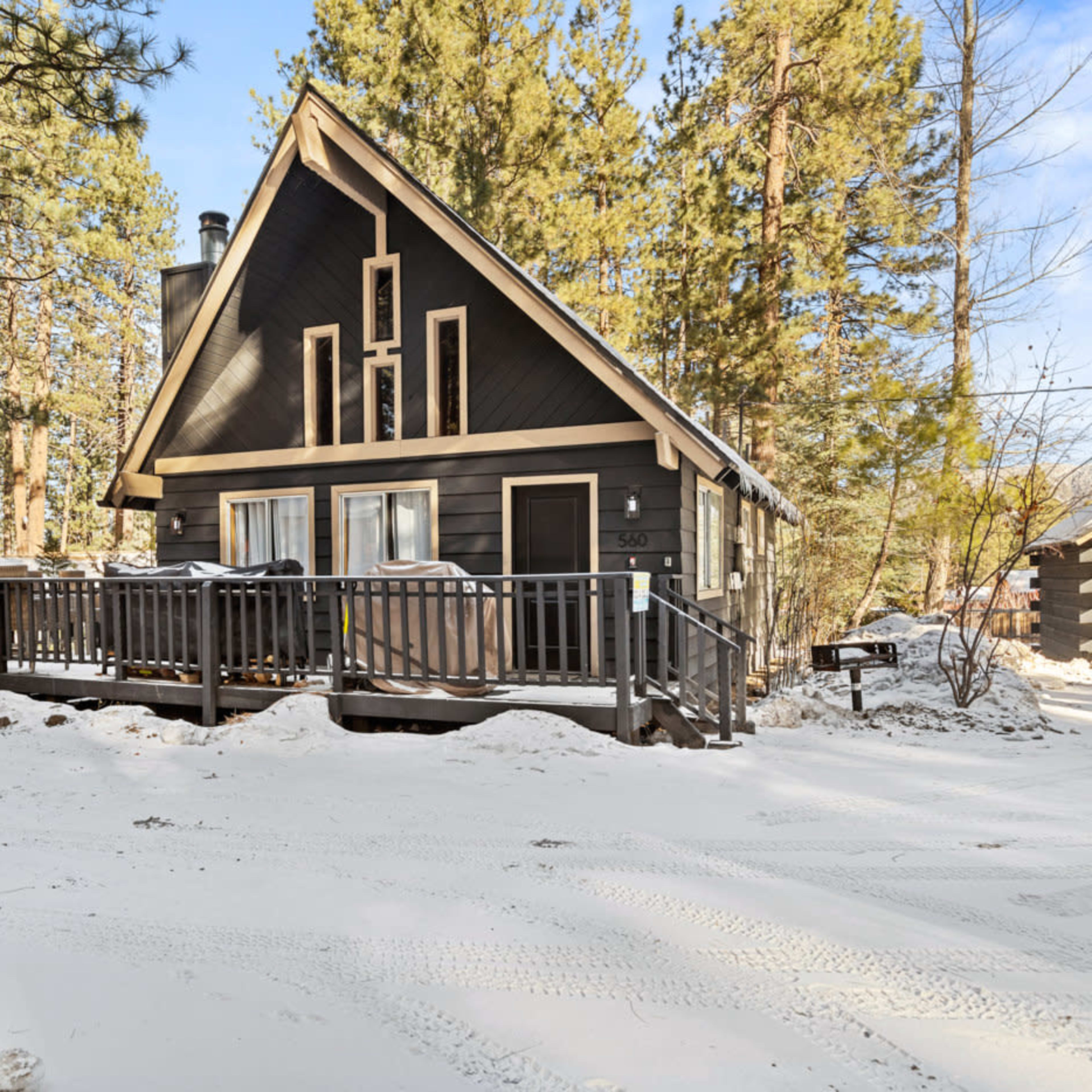 A cabin with a triangular roof and a front deck is situated among tall pine trees in a snowy landscape.