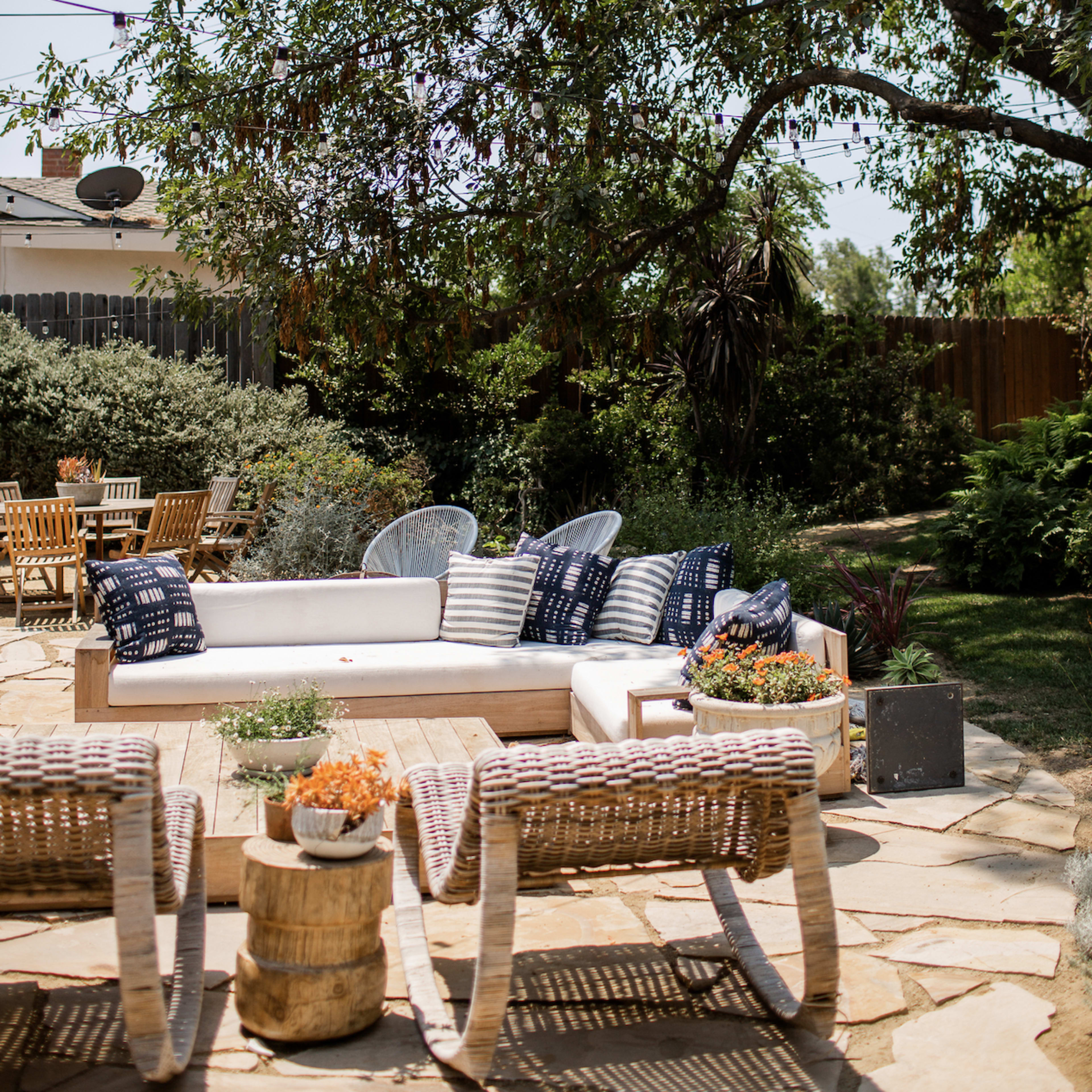 A patio area features a sectional sofa with pillows and a round coffee table surrounded by wicker chairs, all under a large tree in a landscaped outdoor space.