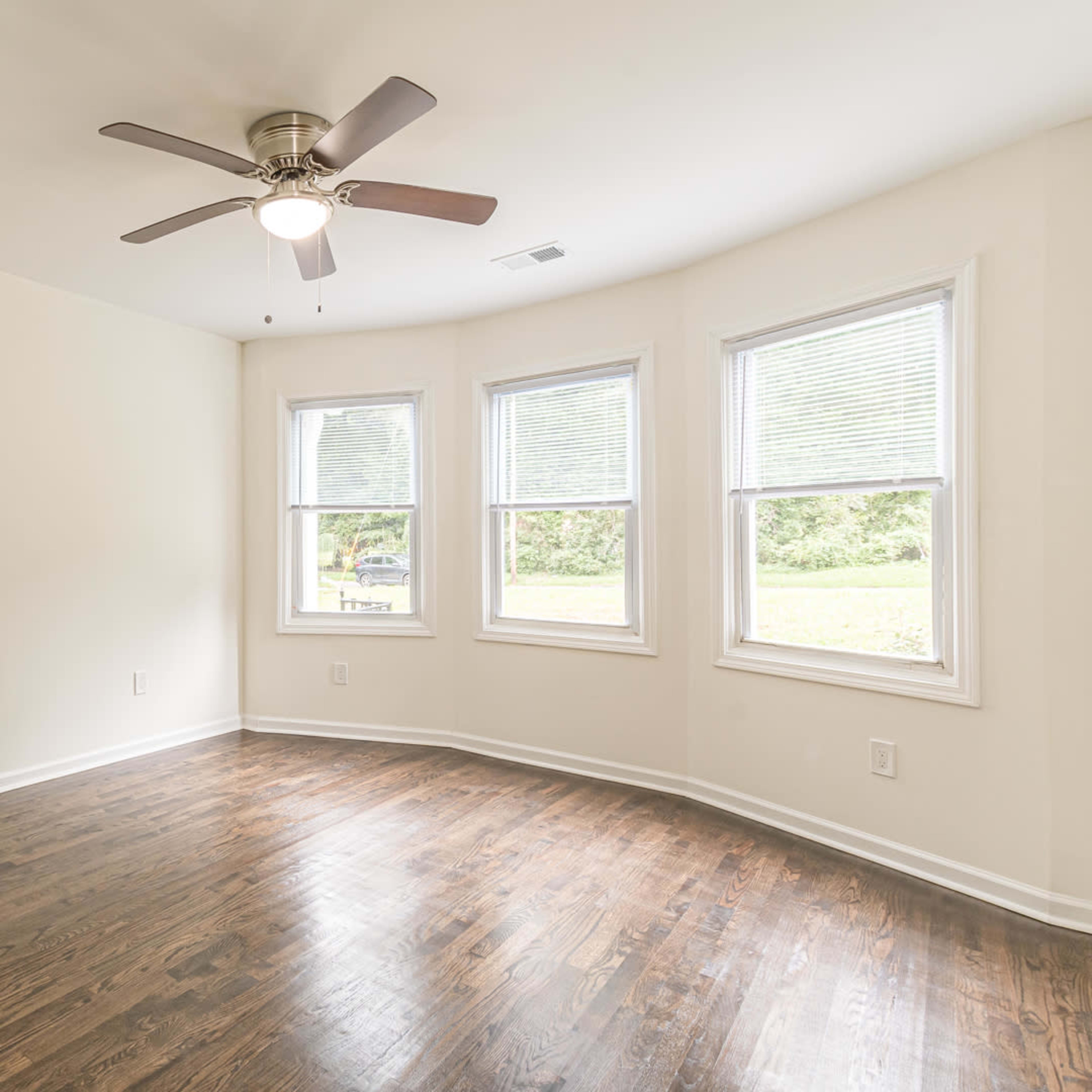 A sparsely furnished room with hardwood floors and three windows allowing natural light.