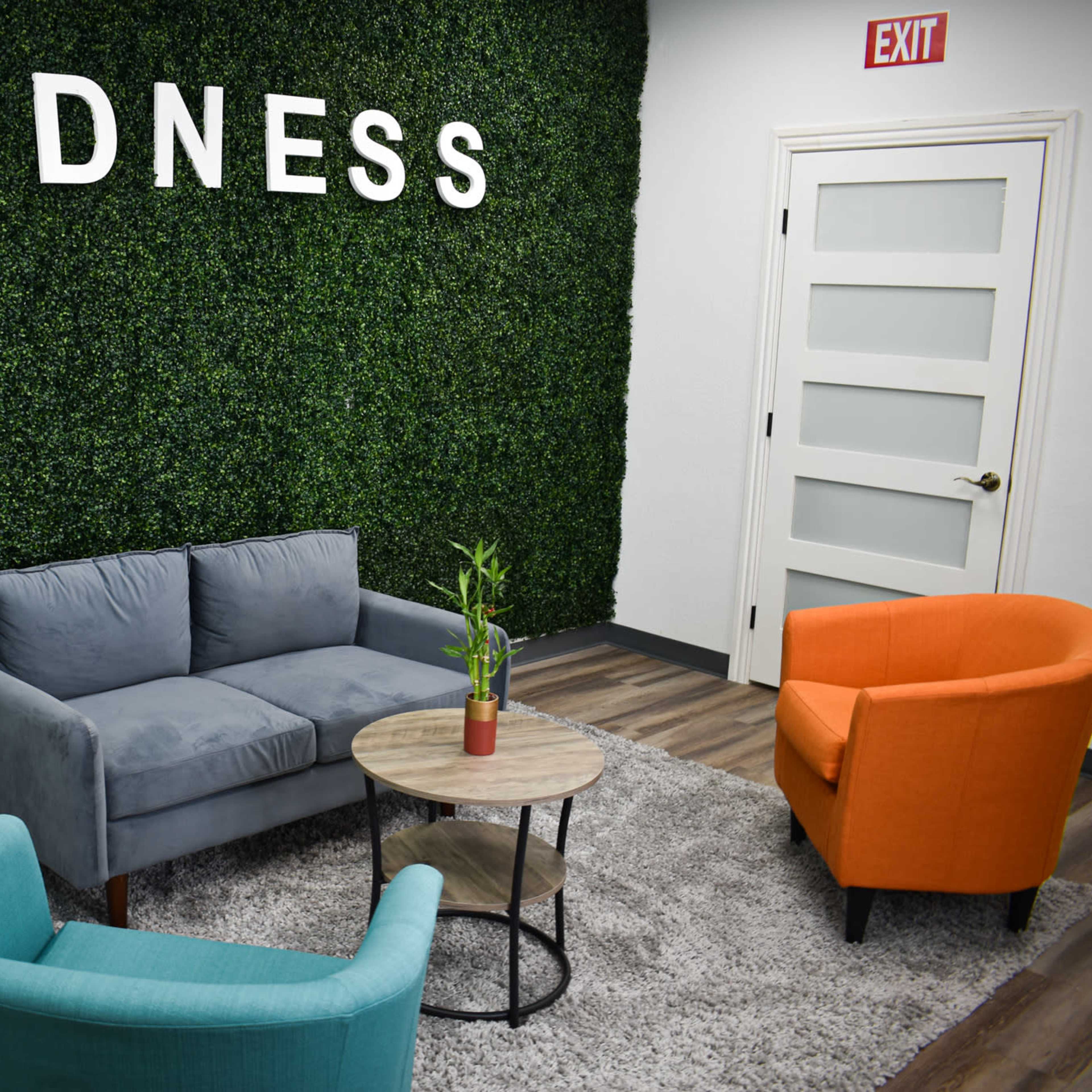 A small seating area with a gray sofa and two colorful chairs in front of a green wall displaying the word "GOODNESS."