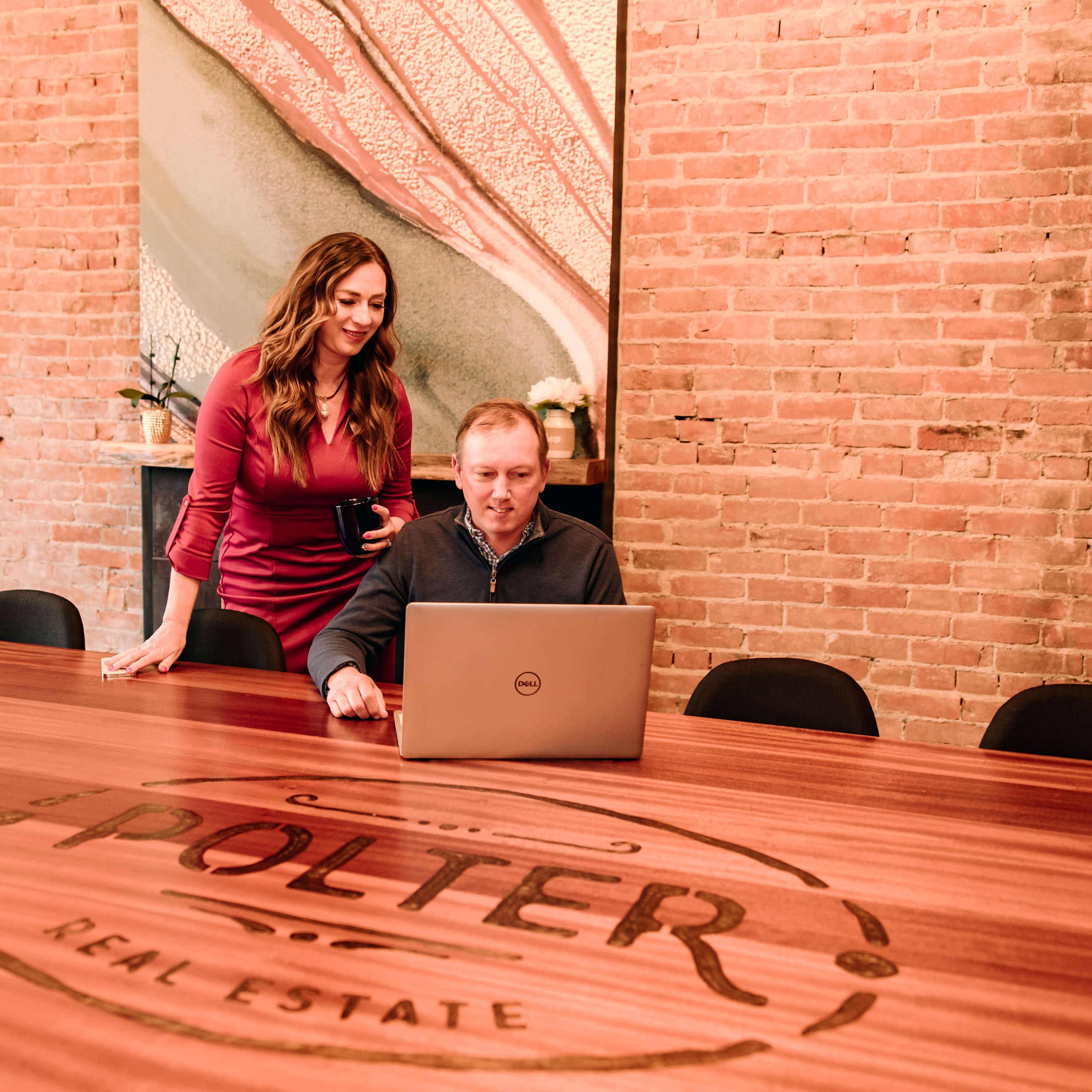 A woman stands beside a man working on a laptop at a wooden conference table marked with "POLTER REAL ESTATE" in a brick-walled meeting room.