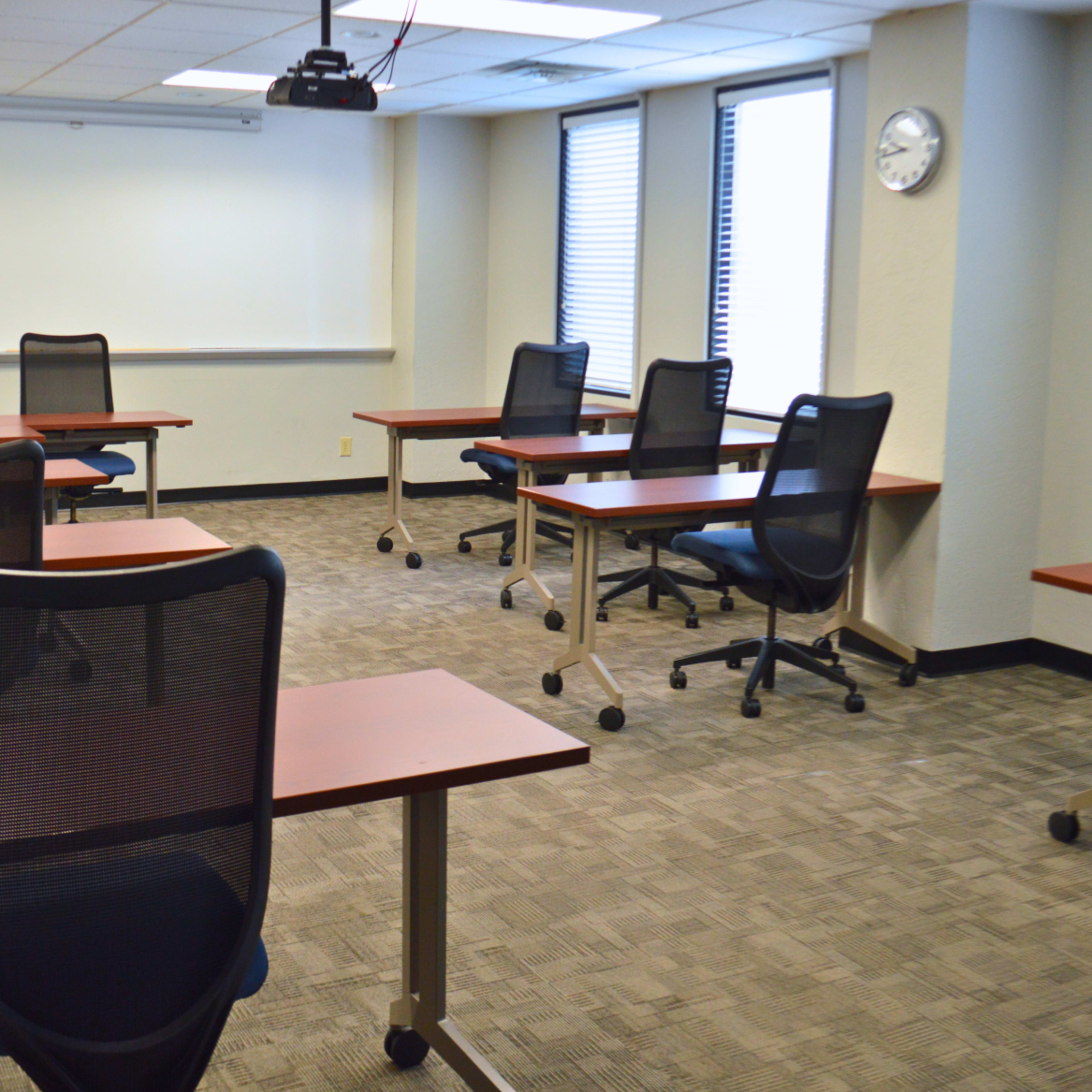 A classroom with several empty desks and chairs arranged in a spacious layout, featuring a whiteboard and a clock on the wall.