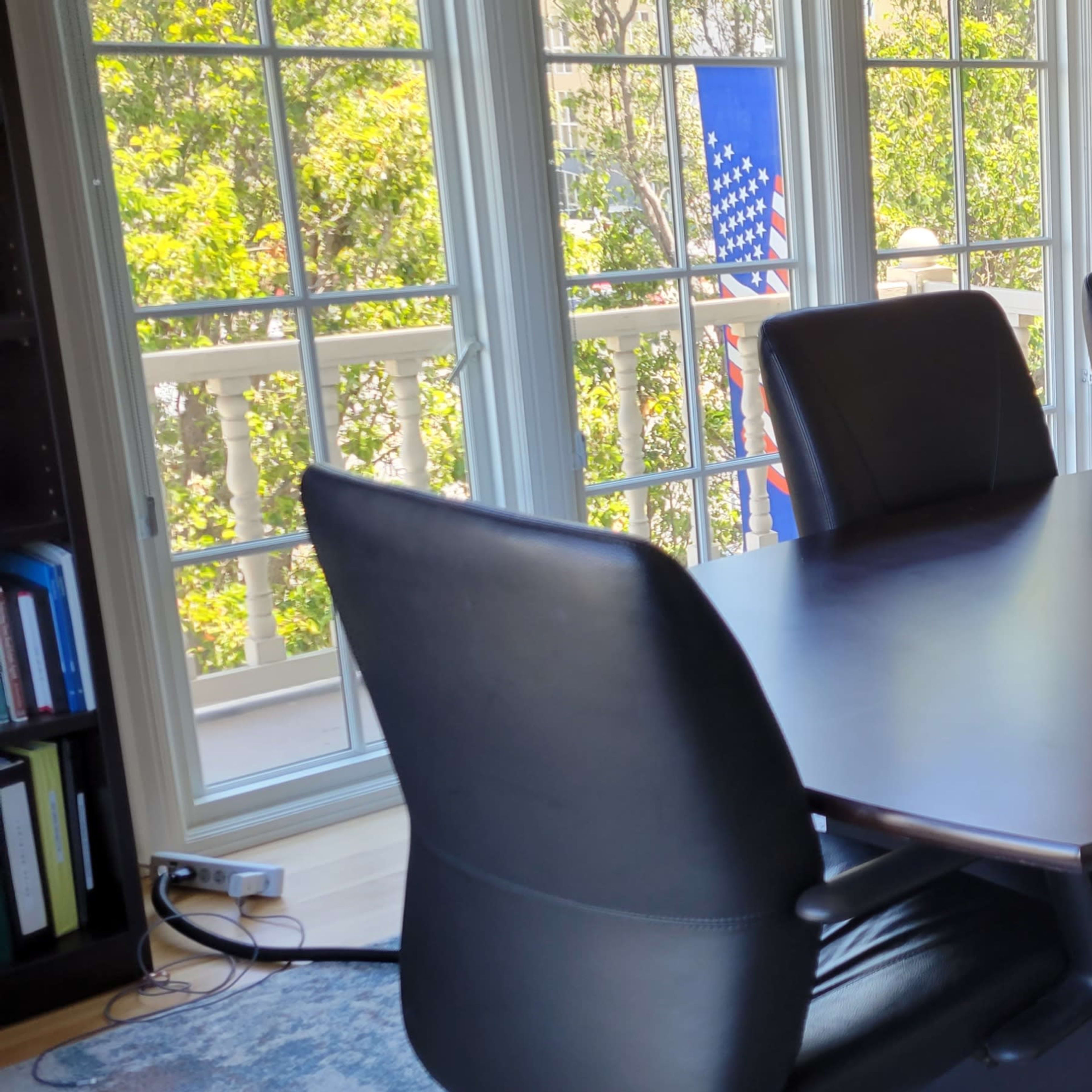 A modern conference room features a large table surrounded by six black ergonomic chairs, with windows displaying an American flag in the background.
