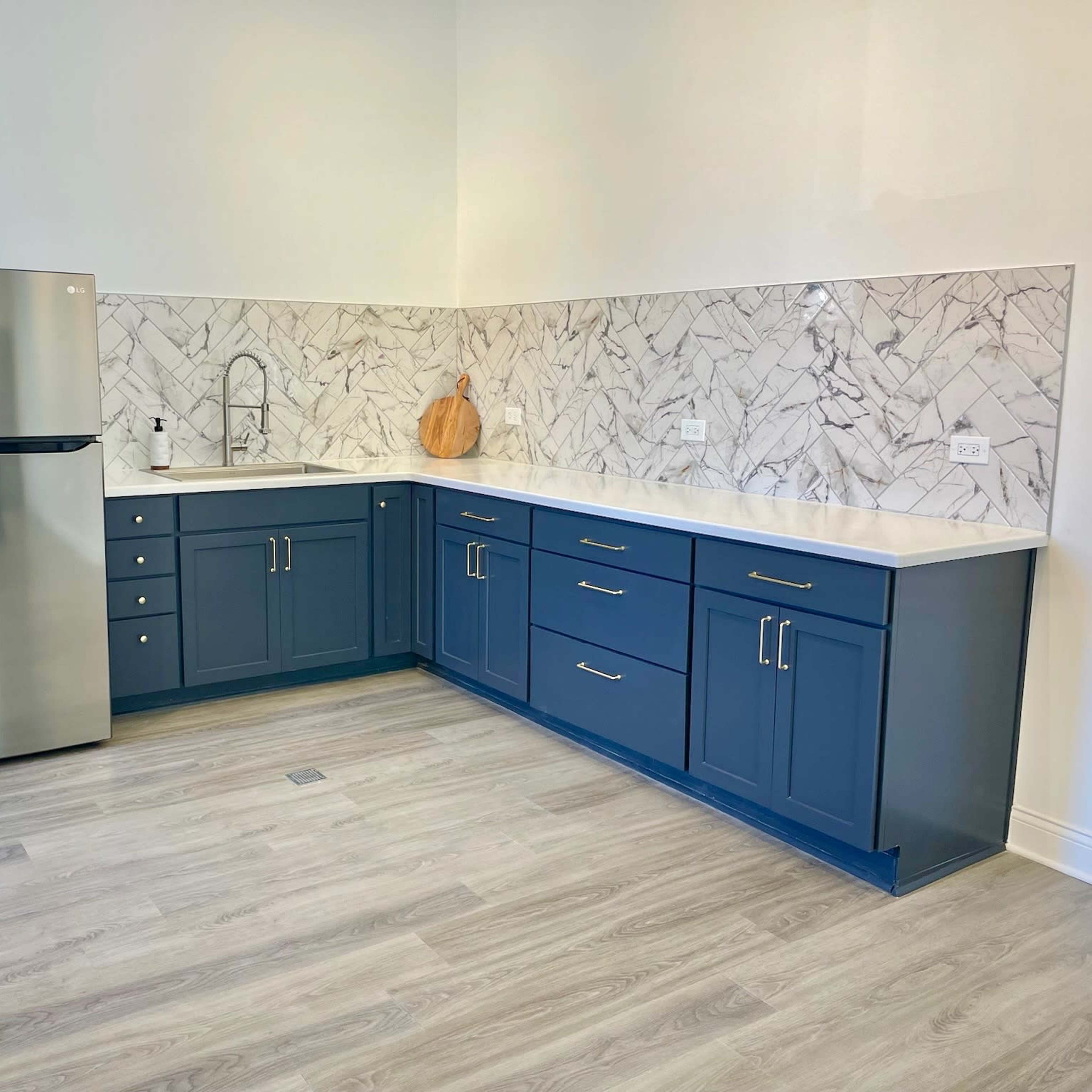 A modern kitchen with blue cabinets, a stainless steel refrigerator, a marble backsplash, and light-colored flooring.