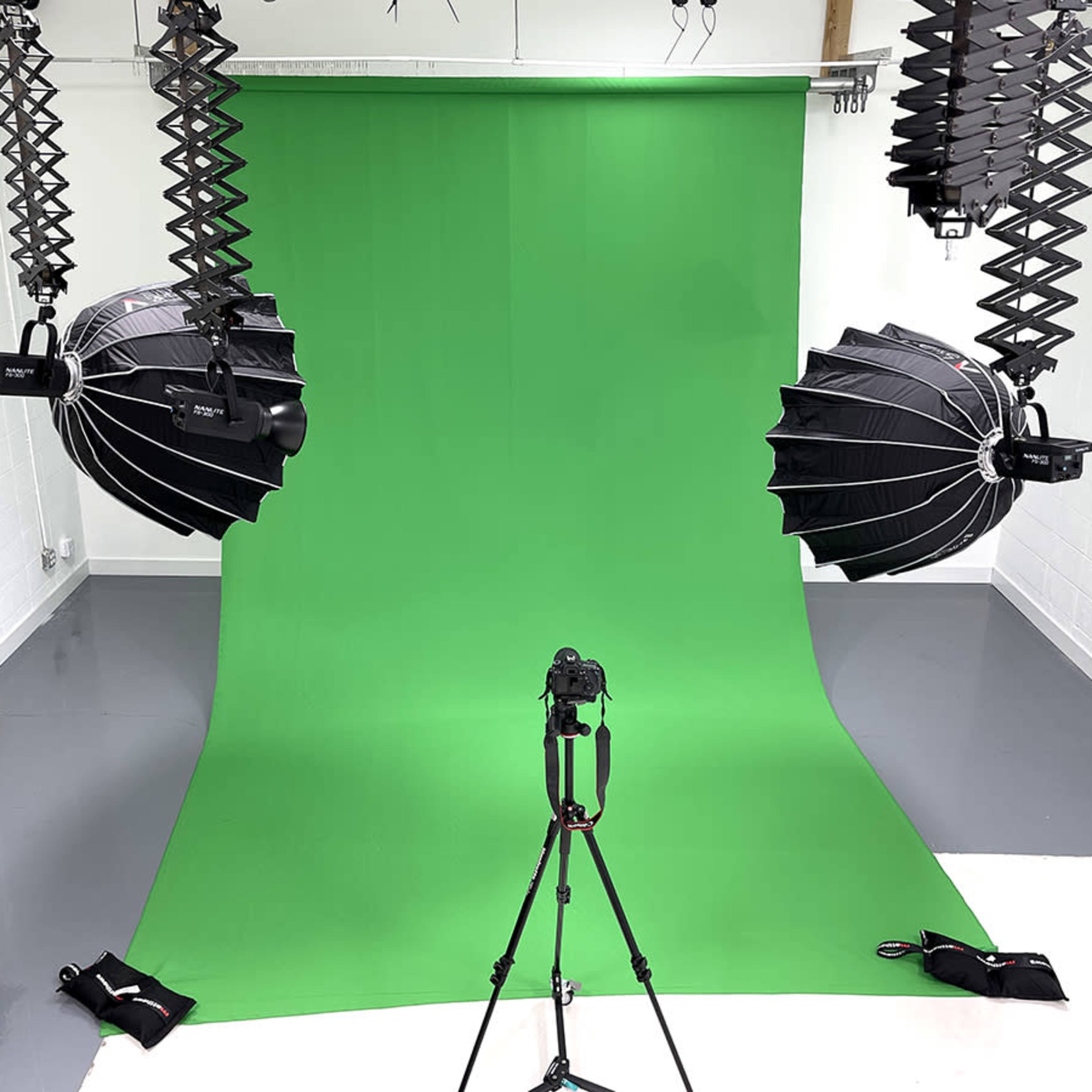 A photography studio with a green backdrop, two overhead lighting fixtures, and a camera mounted on a tripod.