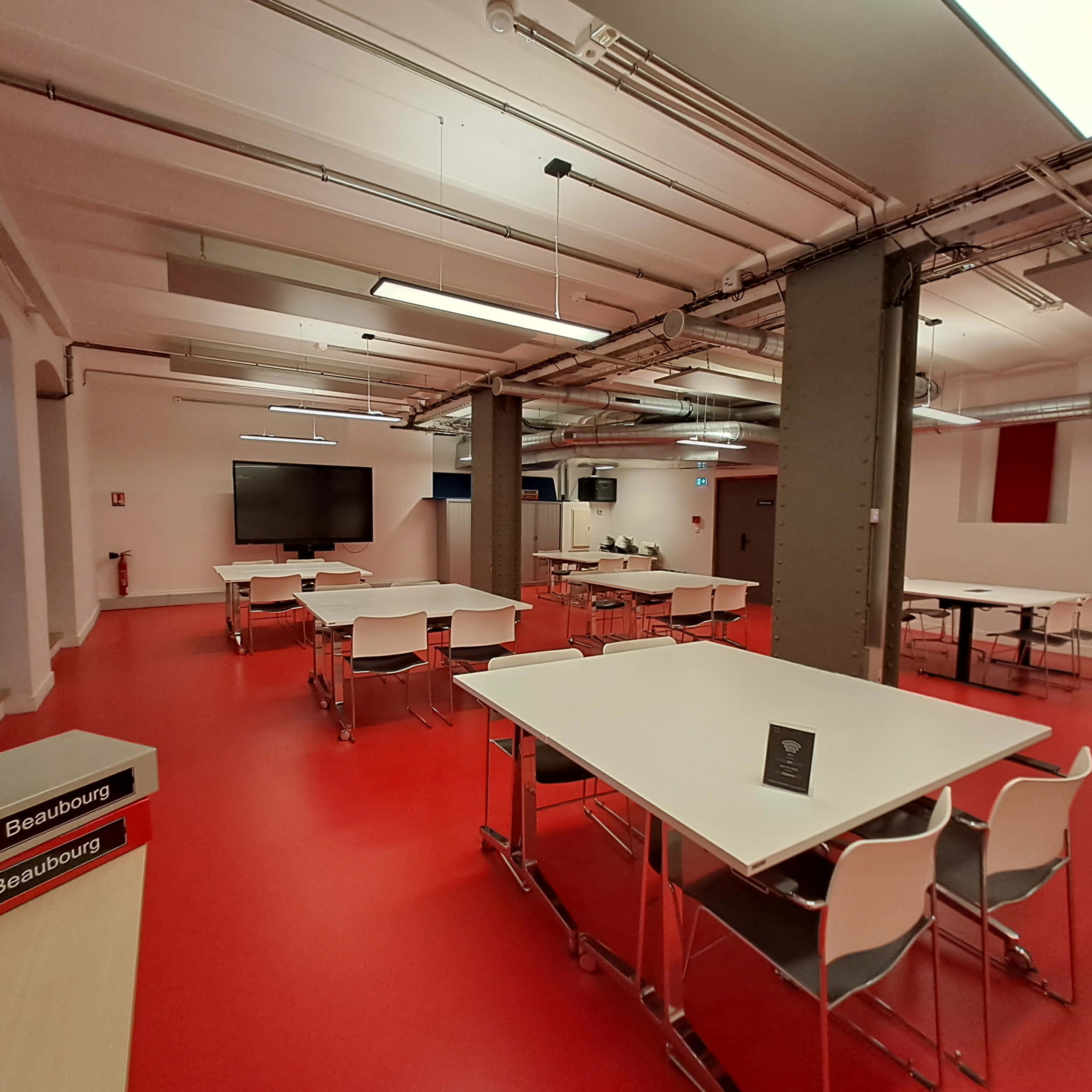 A spacious room with red flooring features multiple white tables and chairs arranged throughout, with a large screen at one end.