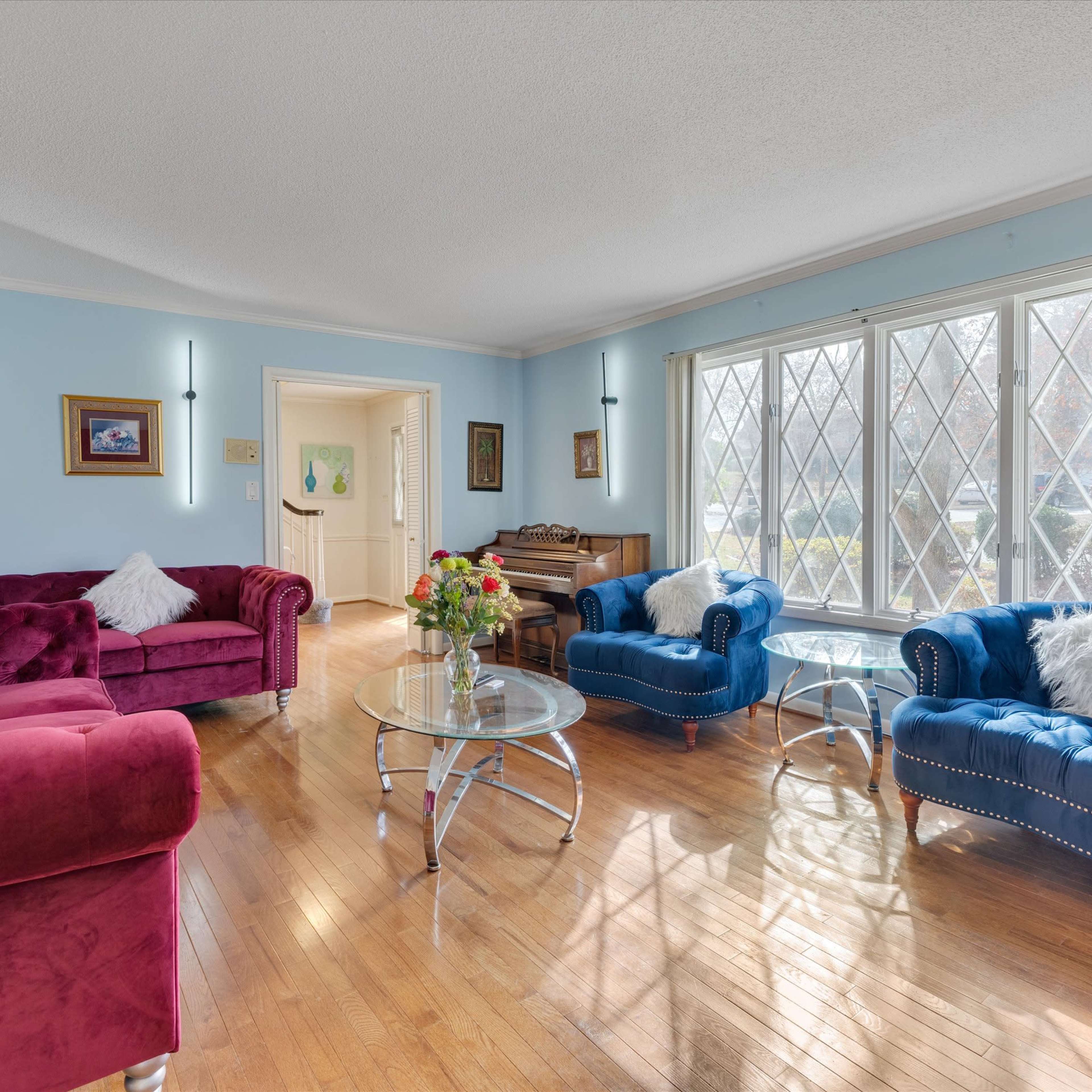 A cozy living room features two upholstered sofas in red and blue, a glass coffee table, and a grand piano near a large bay window.