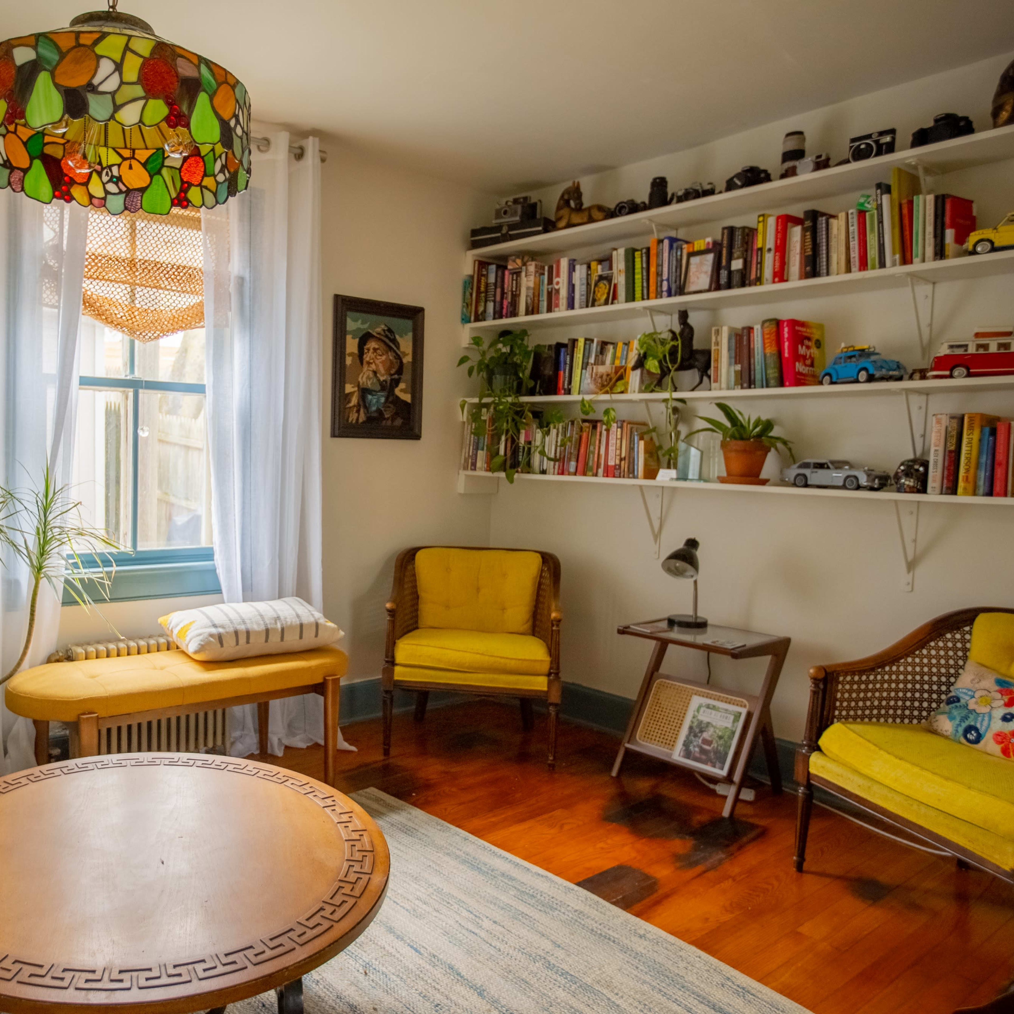 A cozy living room with a round wooden table, yellow chairs, a shelf filled with books and collectibles, and large windows featuring light curtains and indoor plants.