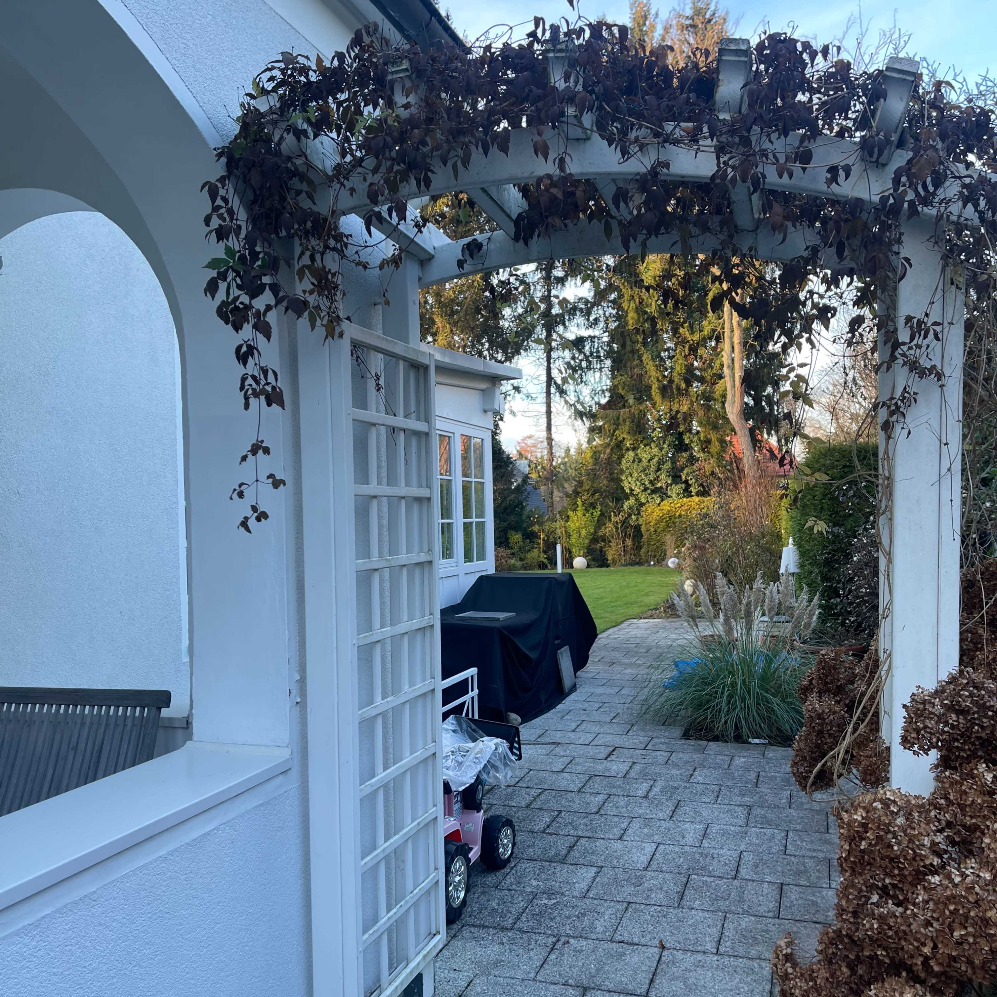 A white trellis arch leads to a paved patio area with a barbecue grill and a garden in the background.