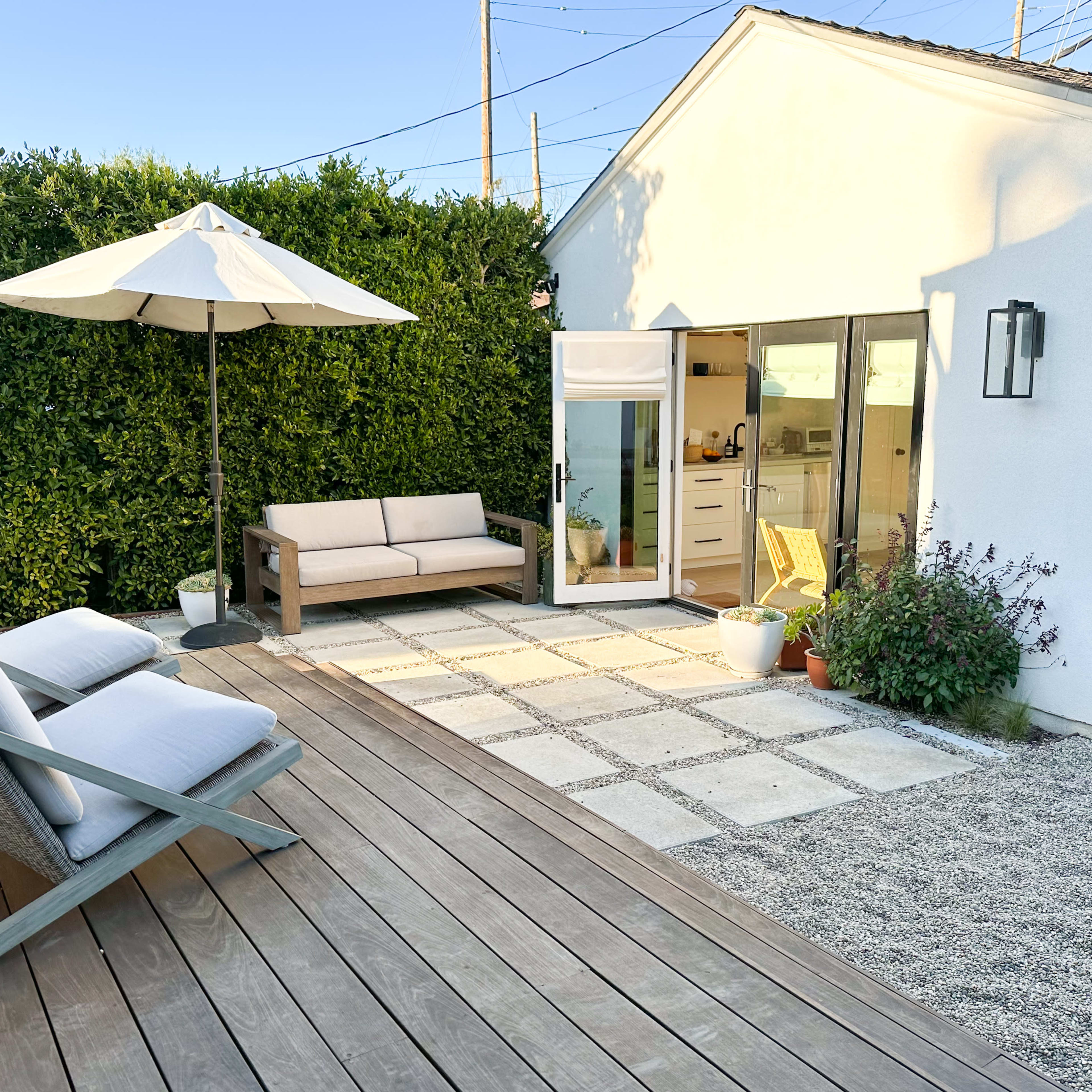 A modern outdoor space features a wooden deck with lounge chairs, a sofa, and a small patio surrounded by greenery and a white house.
