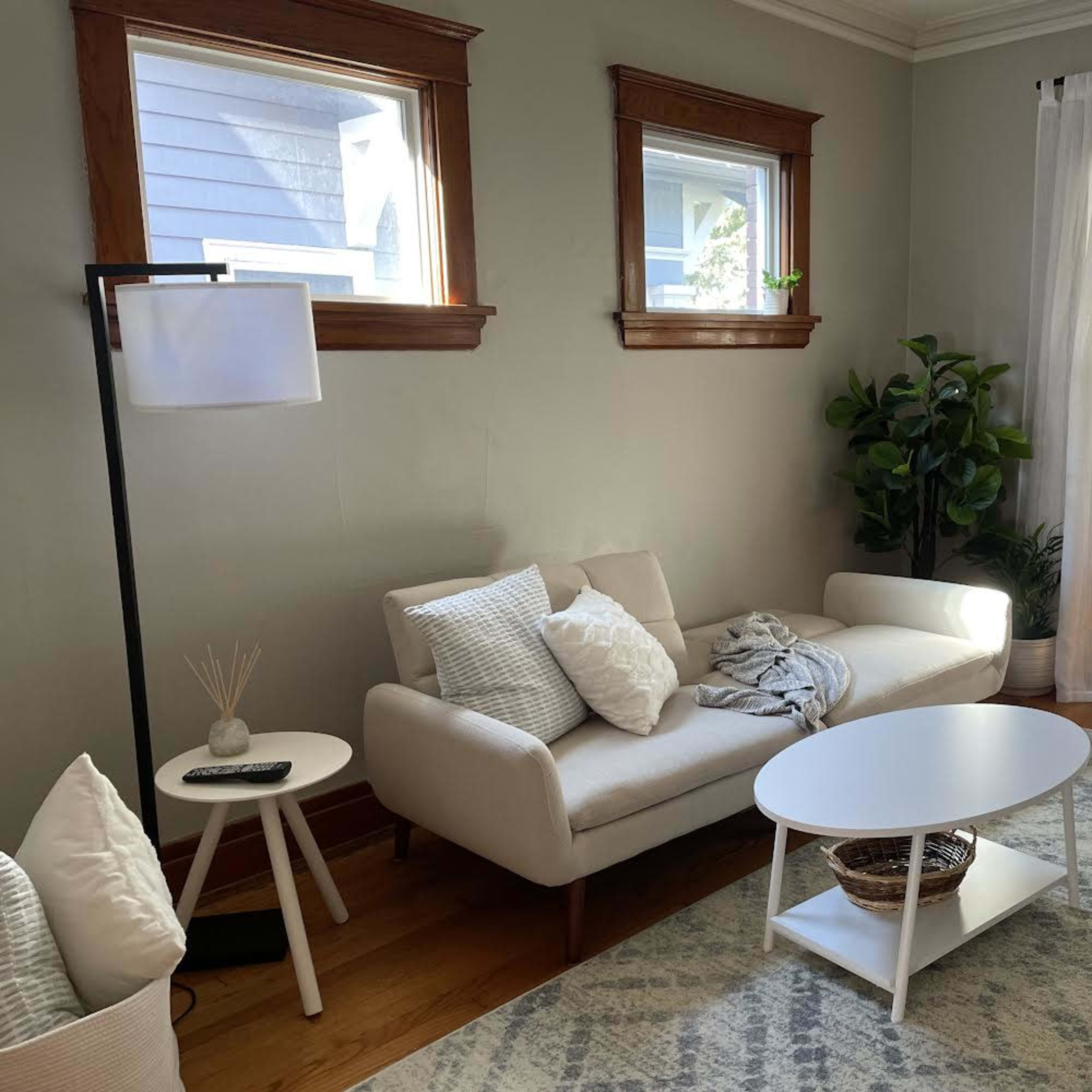 A cozy living room with a white sofa, a small round table, and large windows allowing natural light to enter.