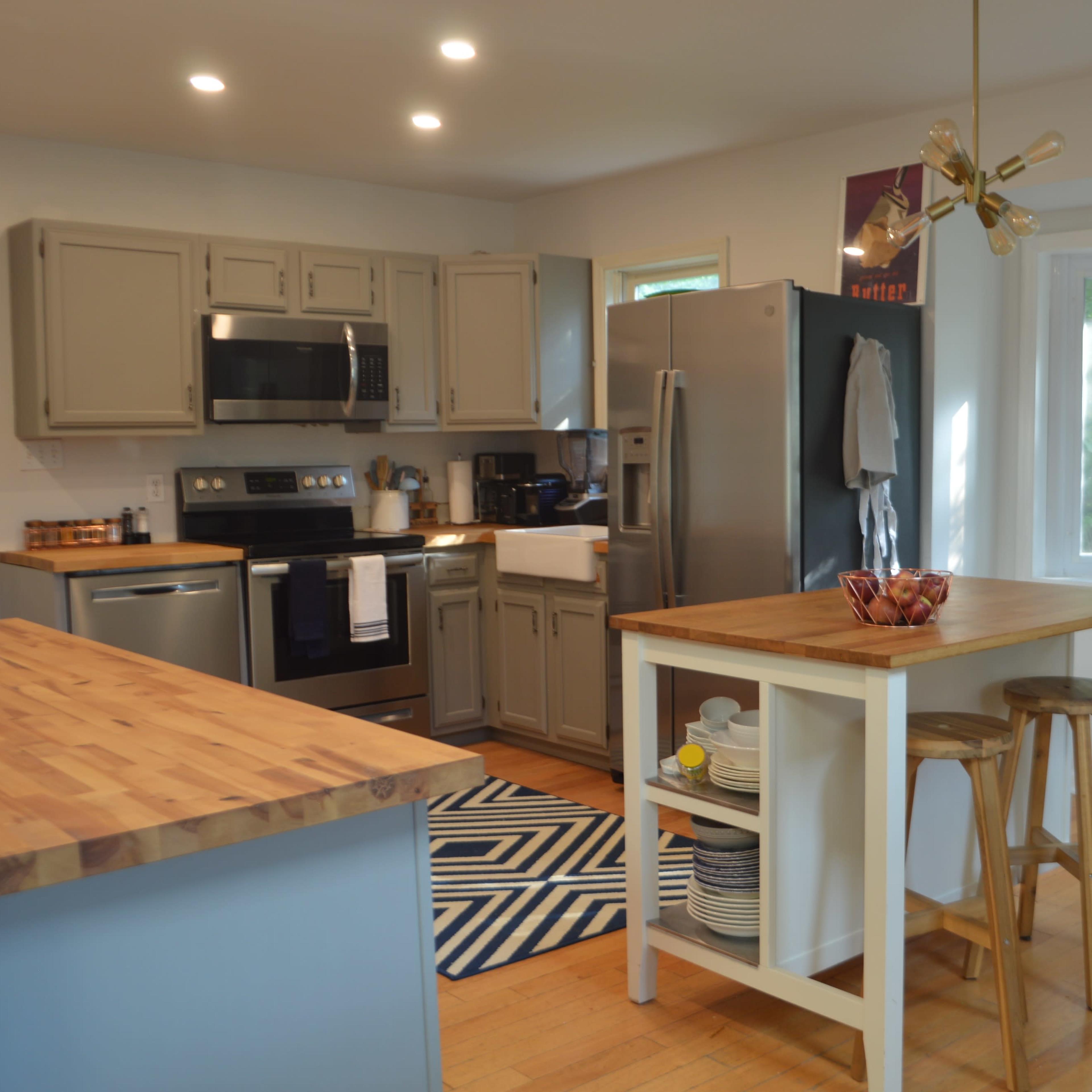 A modern kitchen features grey cabinets, stainless steel appliances, a wooden island with seating, and bright natural light from a nearby window.