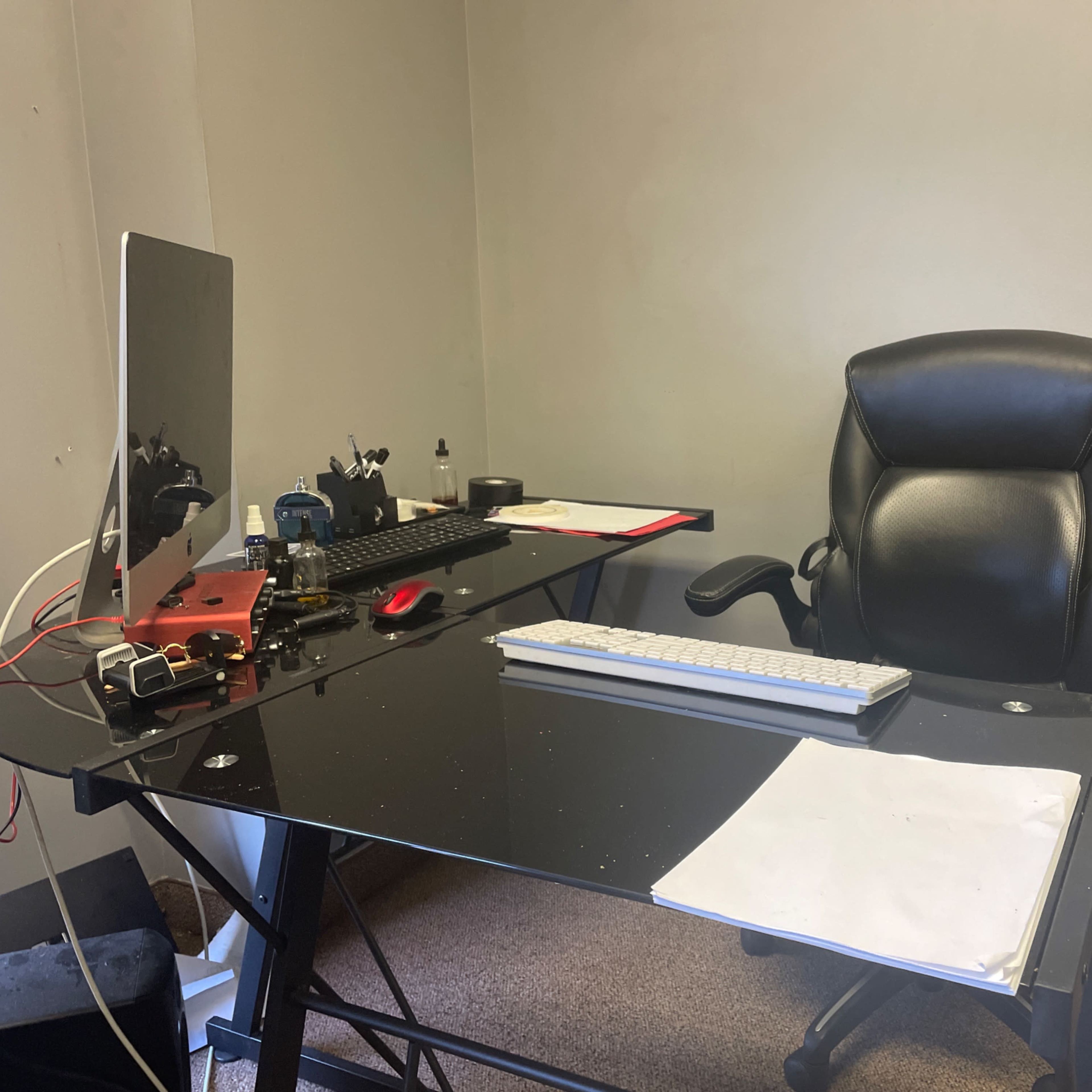 A black office chair next to a glass desk equipped with a computer monitor, keyboard, and some scattered papers.