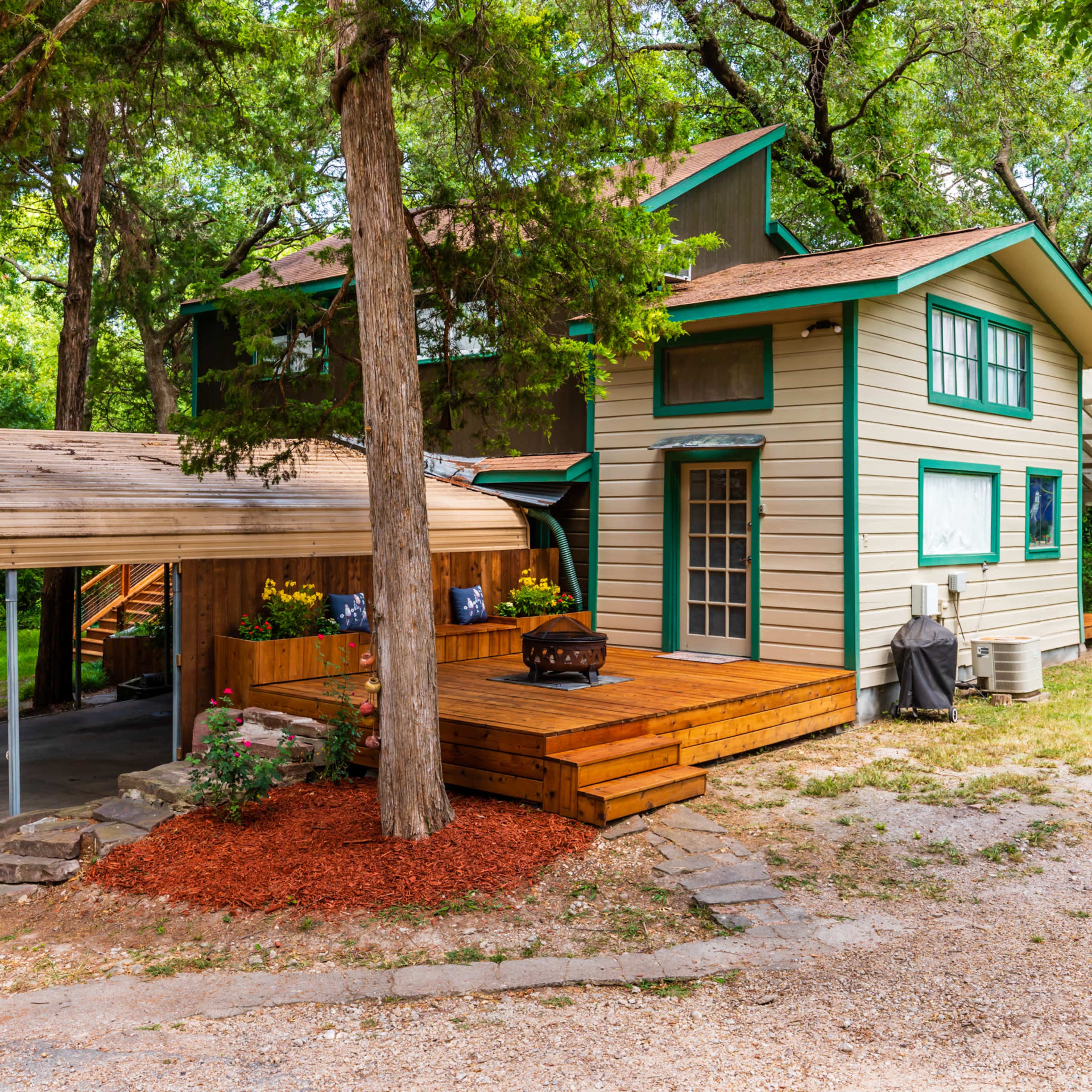 A two-story house with a green and beige exterior features a large wooden deck, a carport, and a landscaped yard surrounded by trees.