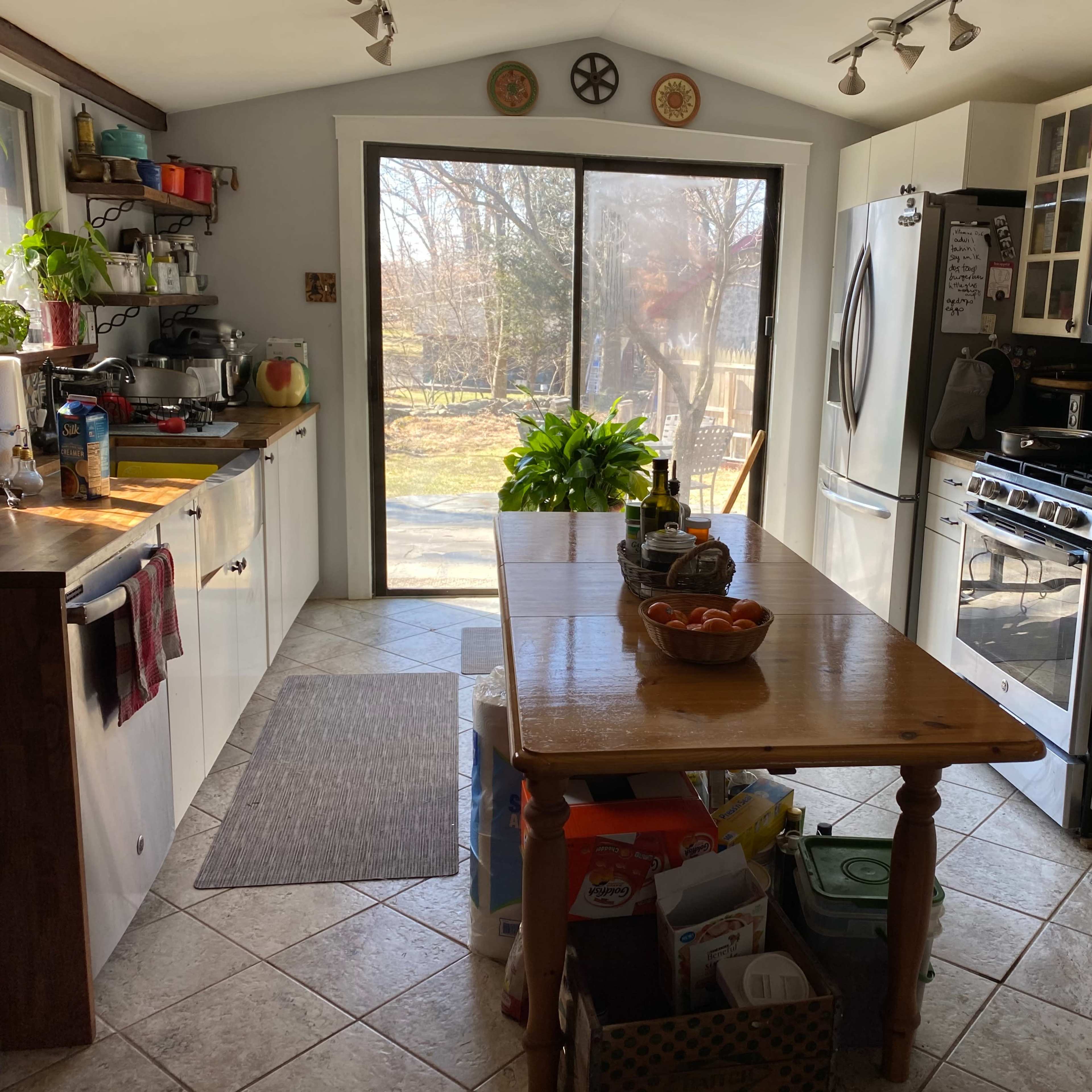 A kitchen with a central wooden table, large windows letting in natural light, and various appliances and plants visible.