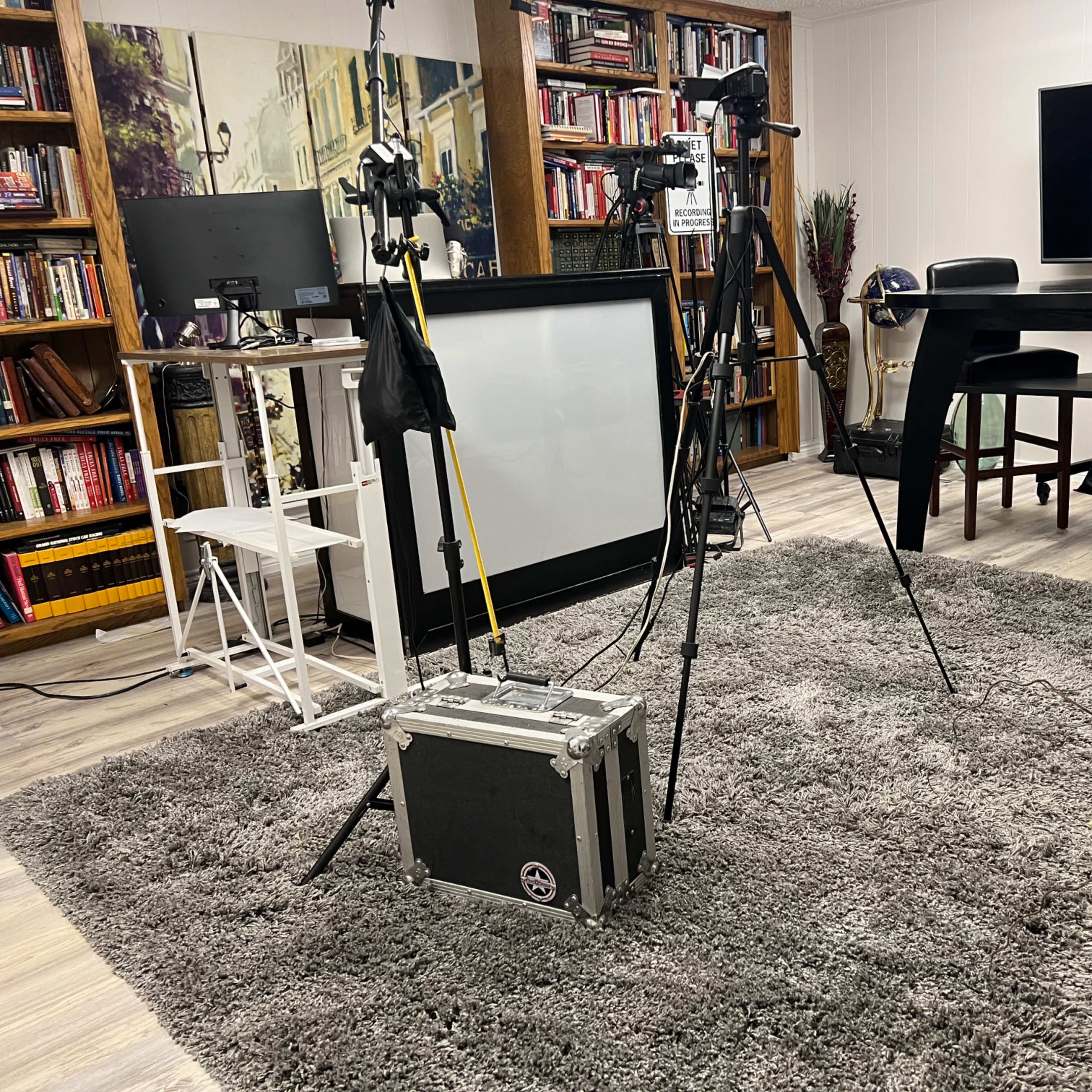 A room set up for a video shoot, featuring tripods, a monitor, a black table, and a large grey rug.