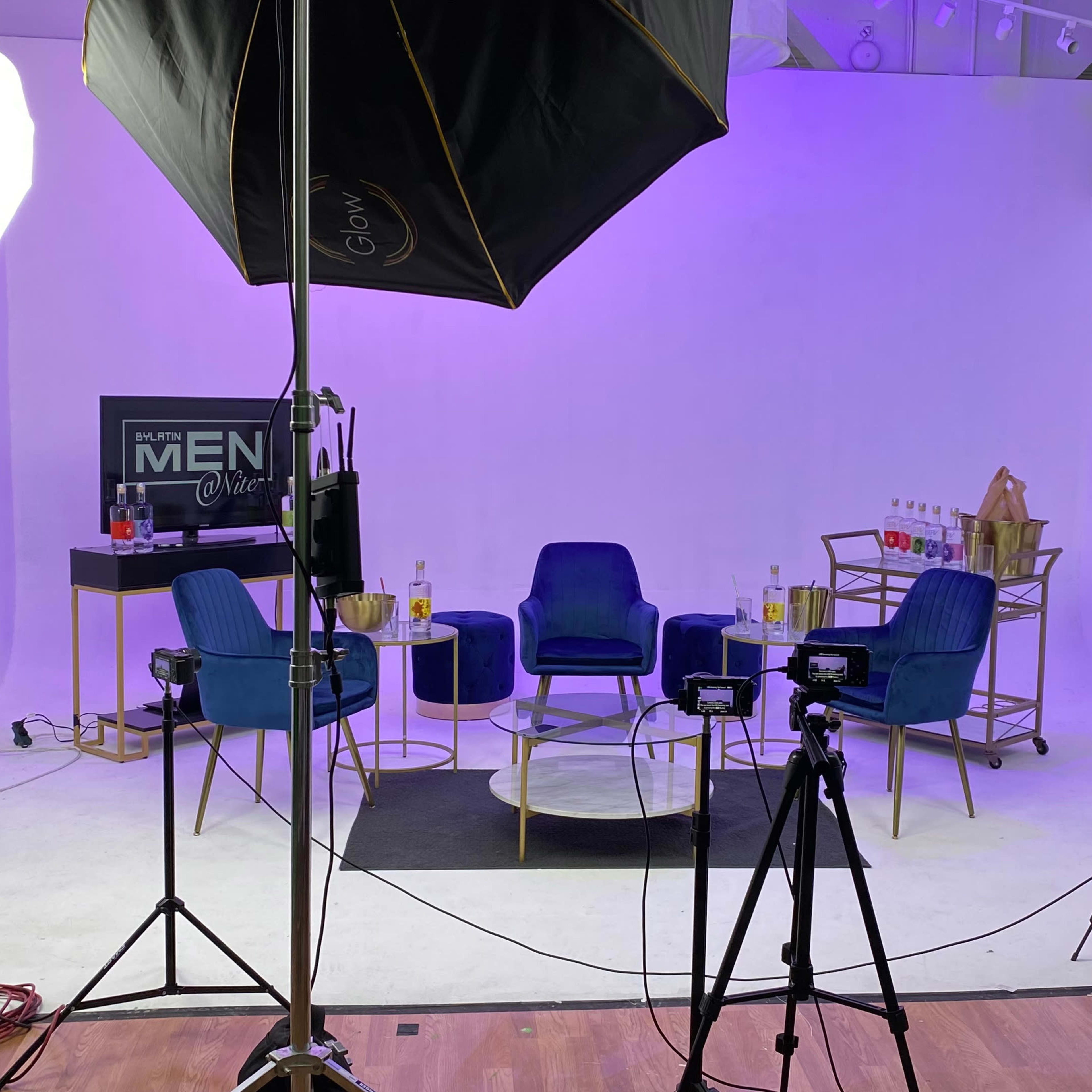 A studio setup with four blue chairs arranged around a glass coffee table, with lighting equipment and cameras positioned nearby.