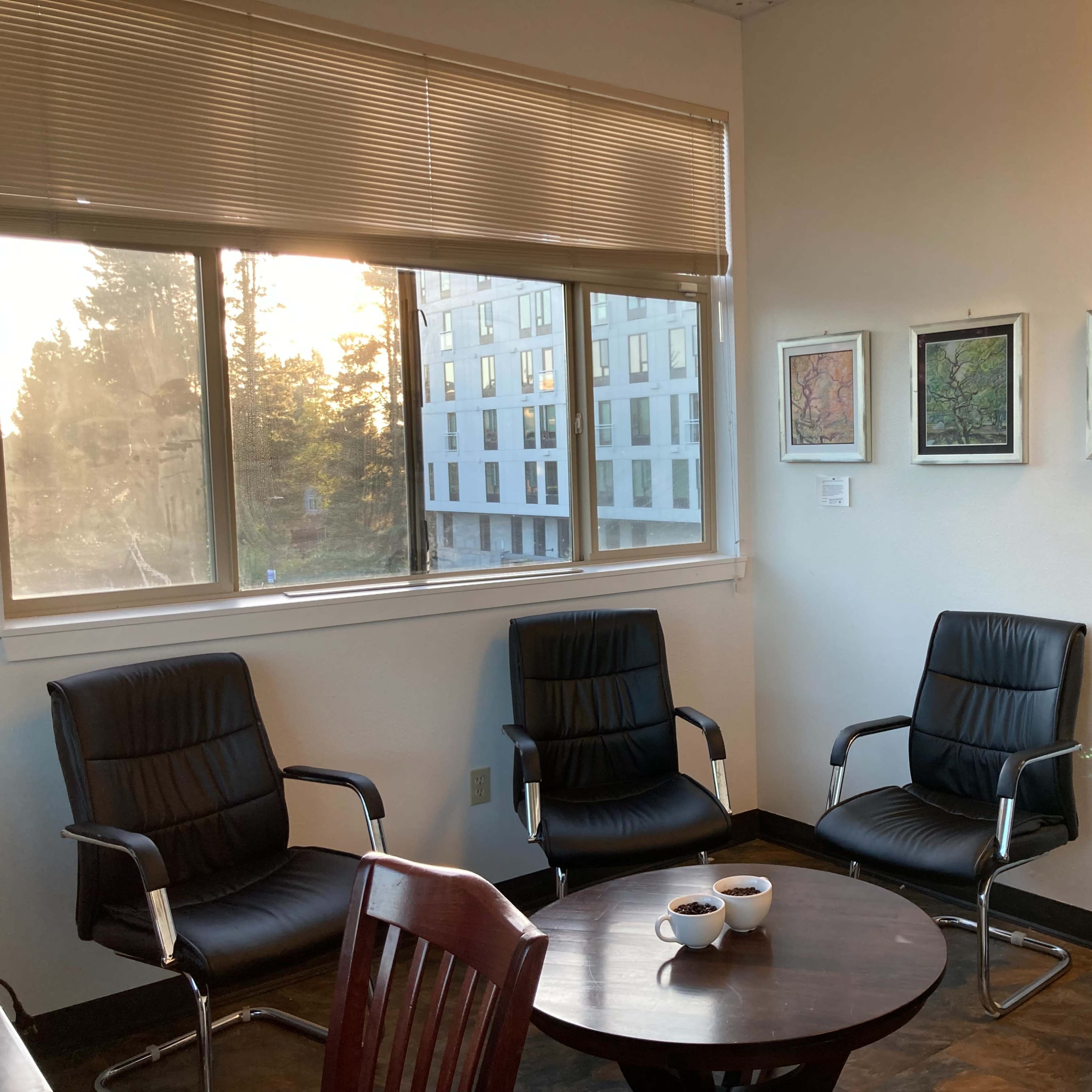 A waiting room features four black chairs surrounding a round table with two cups, and colorful artwork displayed on the walls near a window.