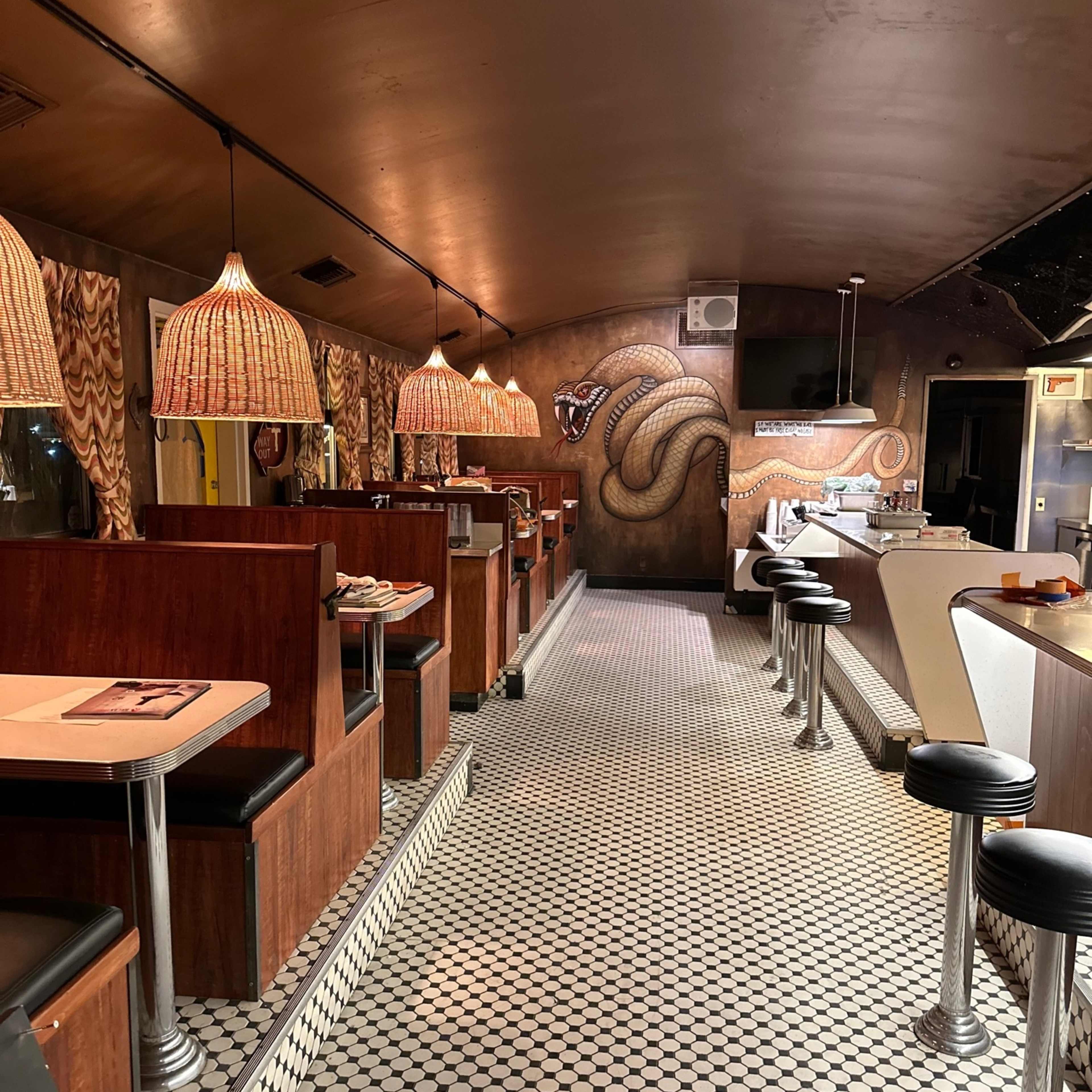 The interior of a retro-style diner with wooden booths, woven pendant lights, and a tiled floor featuring black and white patterns.