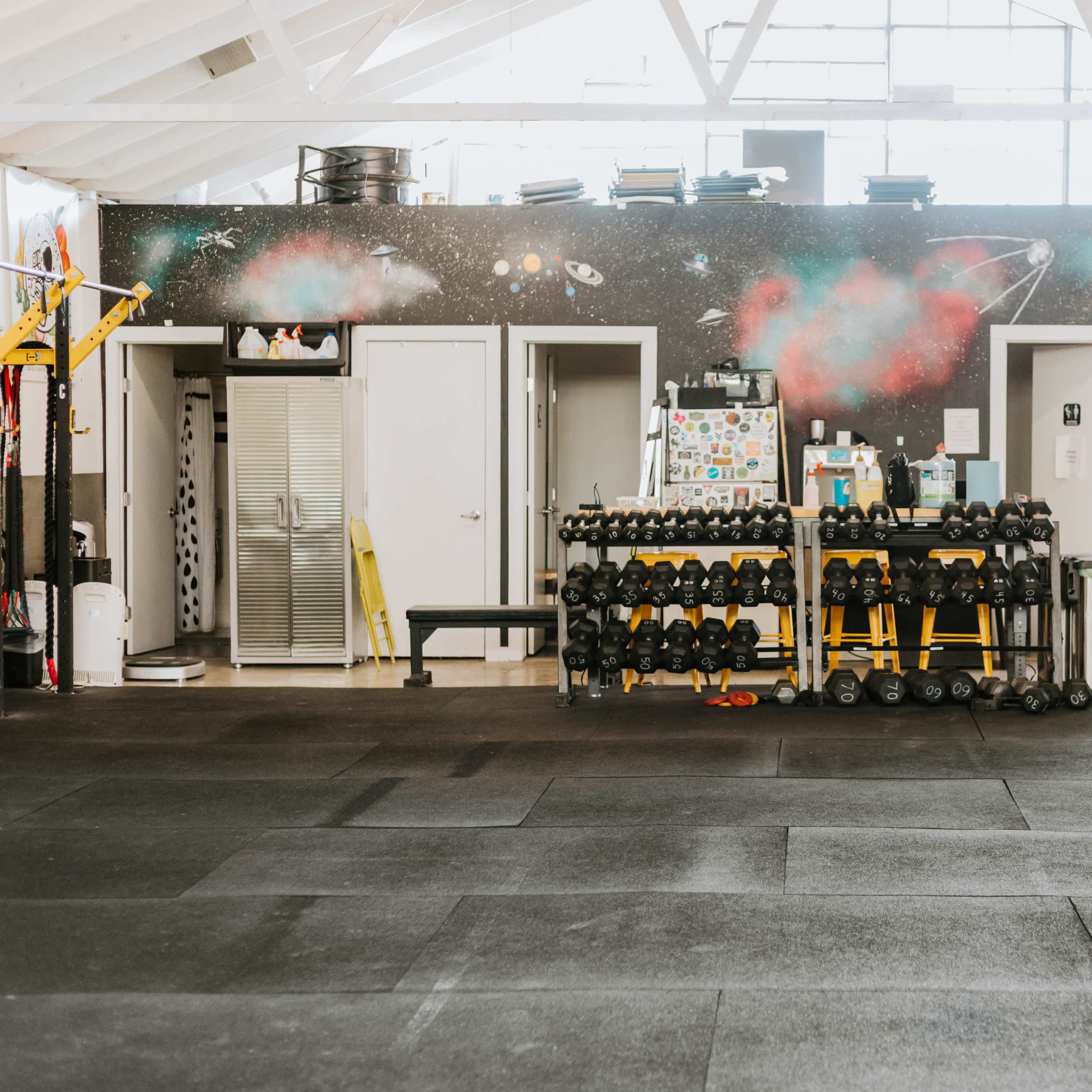 A gym interior with rubber flooring, a weight rack filled with dumbbells, and lockers against a wall featuring a space-themed mural.