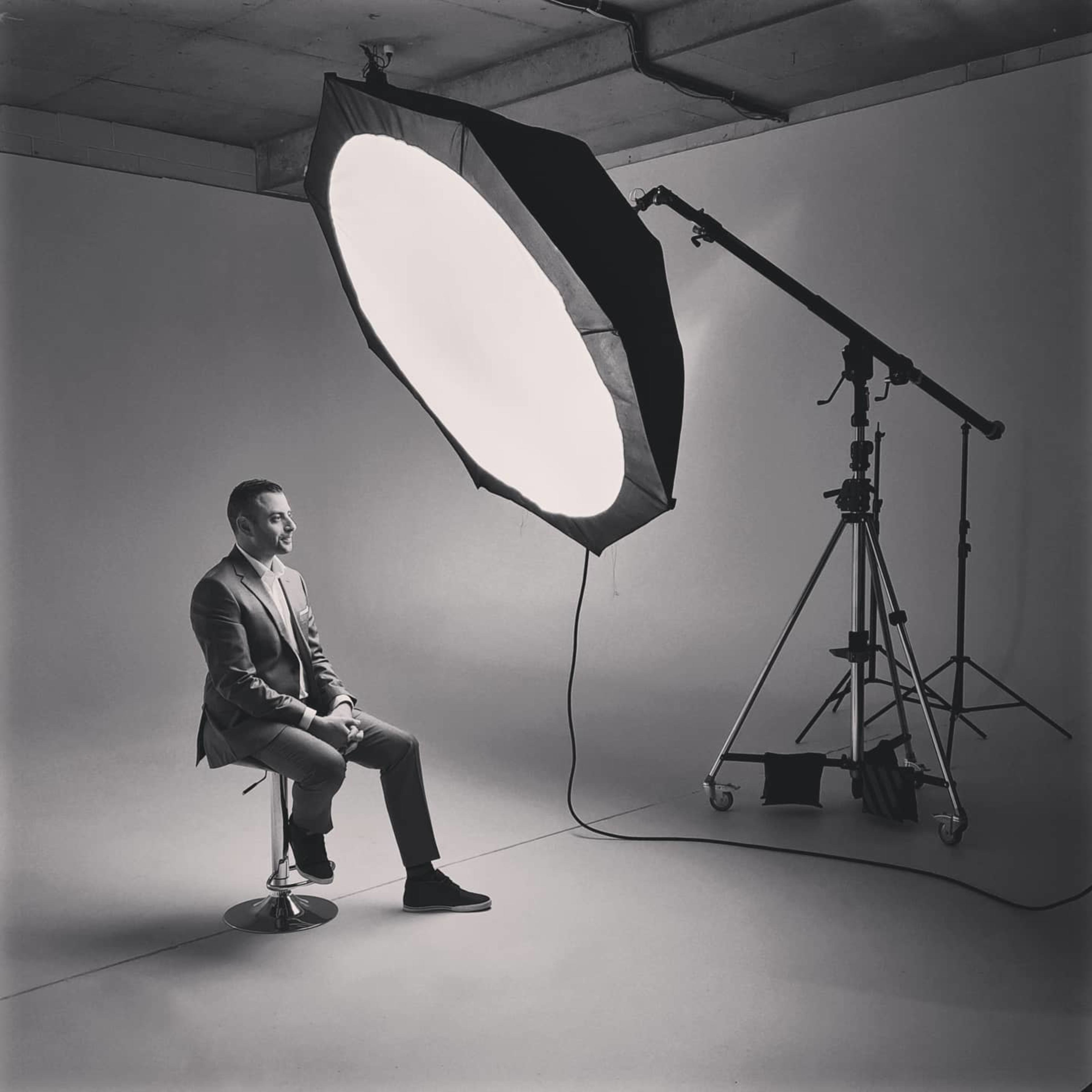 A man in a suit sits on a stool in front of a large, round softbox light in a photography studio.