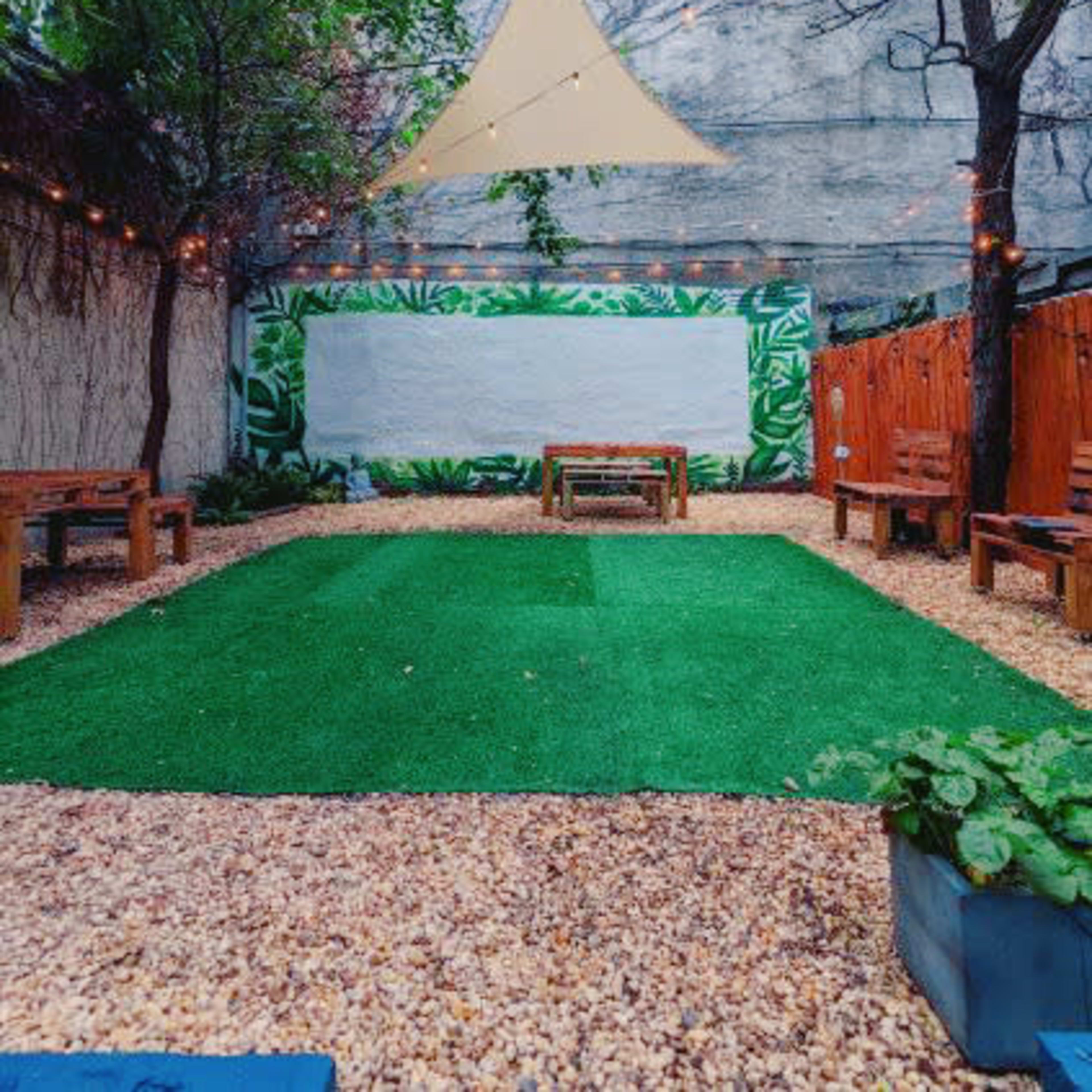 A small outdoor space with artificial grass, wooden benches, and a mural of greenery along one wall, all under a triangular canopy.