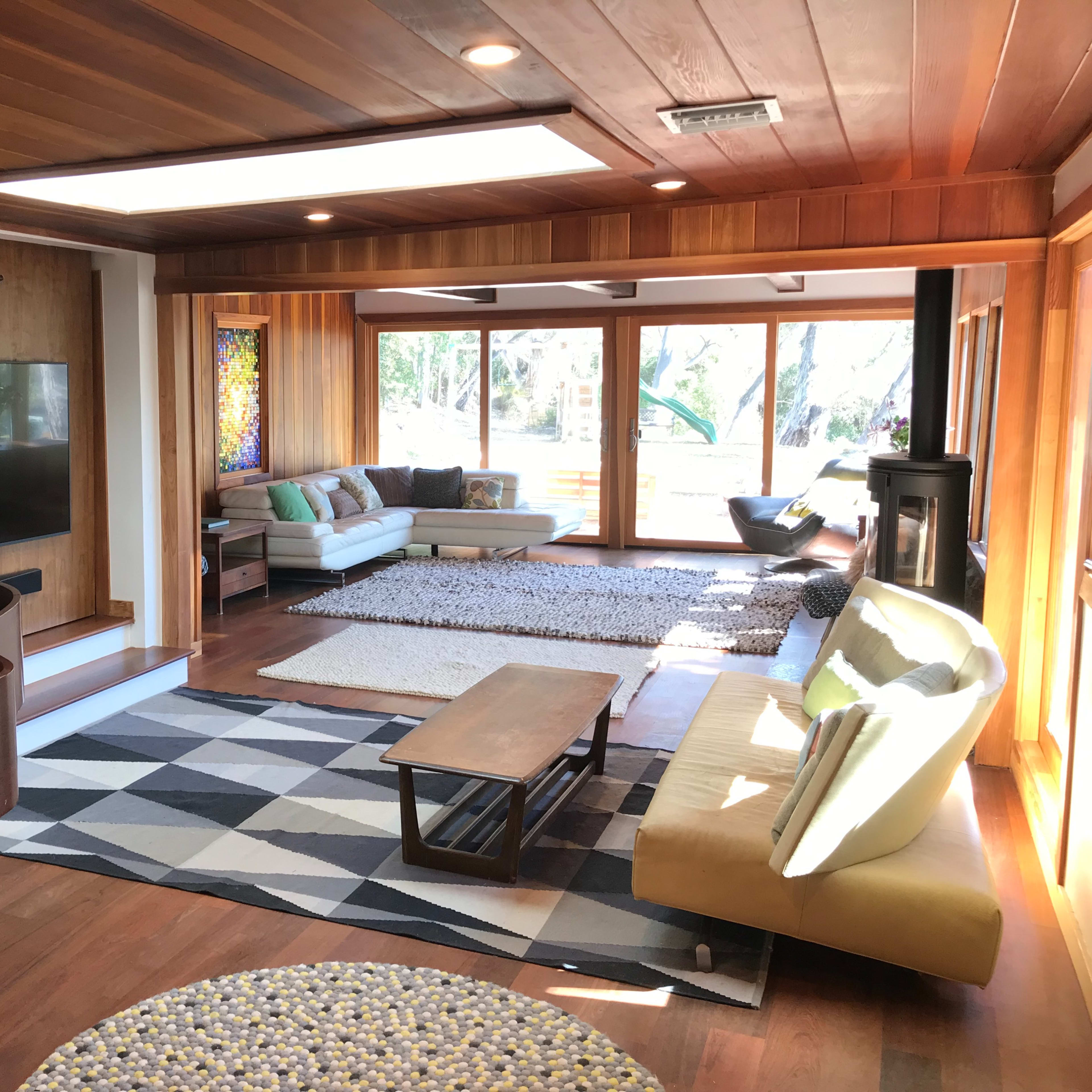 The scene shows a spacious living room with wooden paneling, a grand piano, a modern sofa arrangement, and large windows overlooking a landscaped outdoor area.