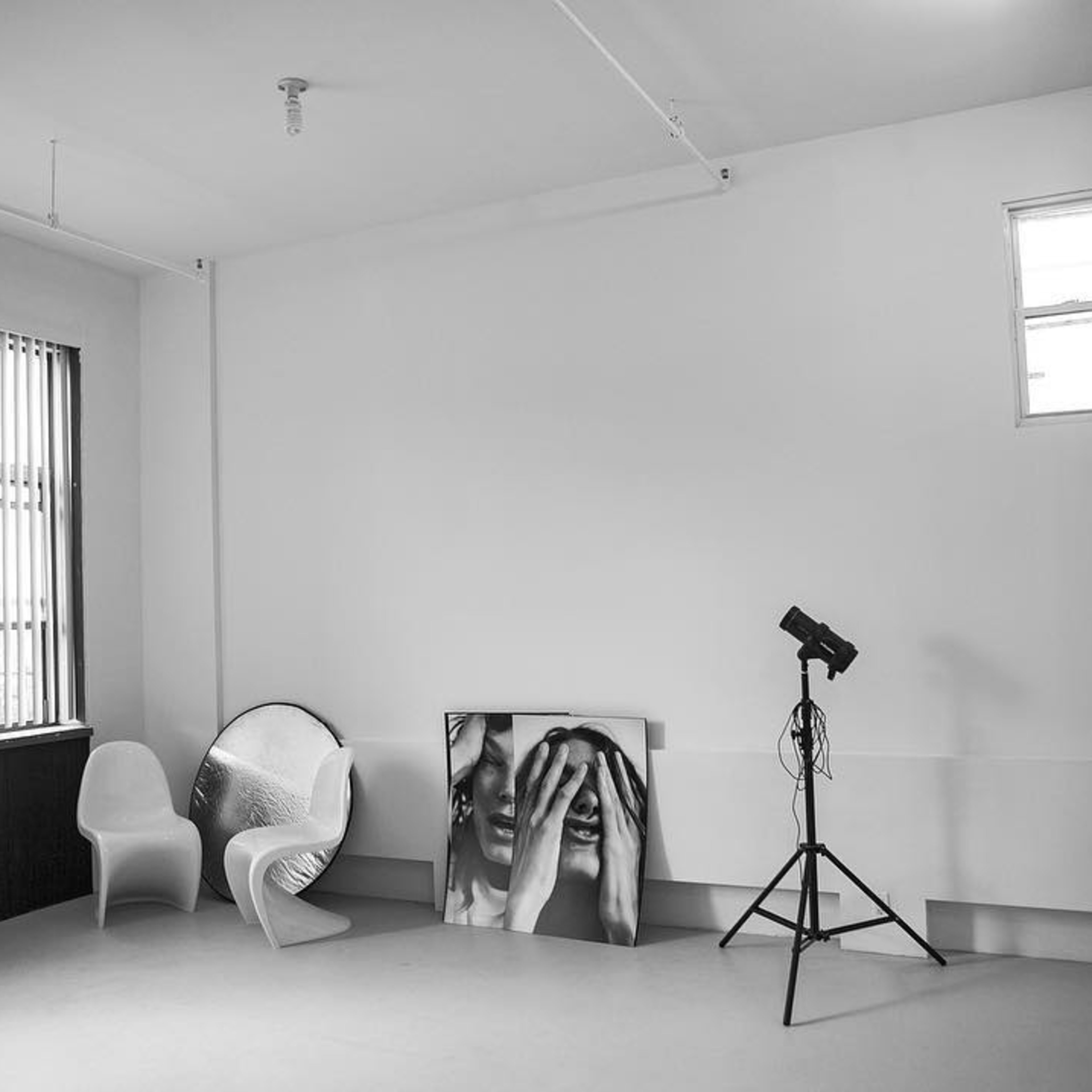 A minimalist studio space with two chairs, a framed black-and-white photograph, and a lighting stand near a window.
