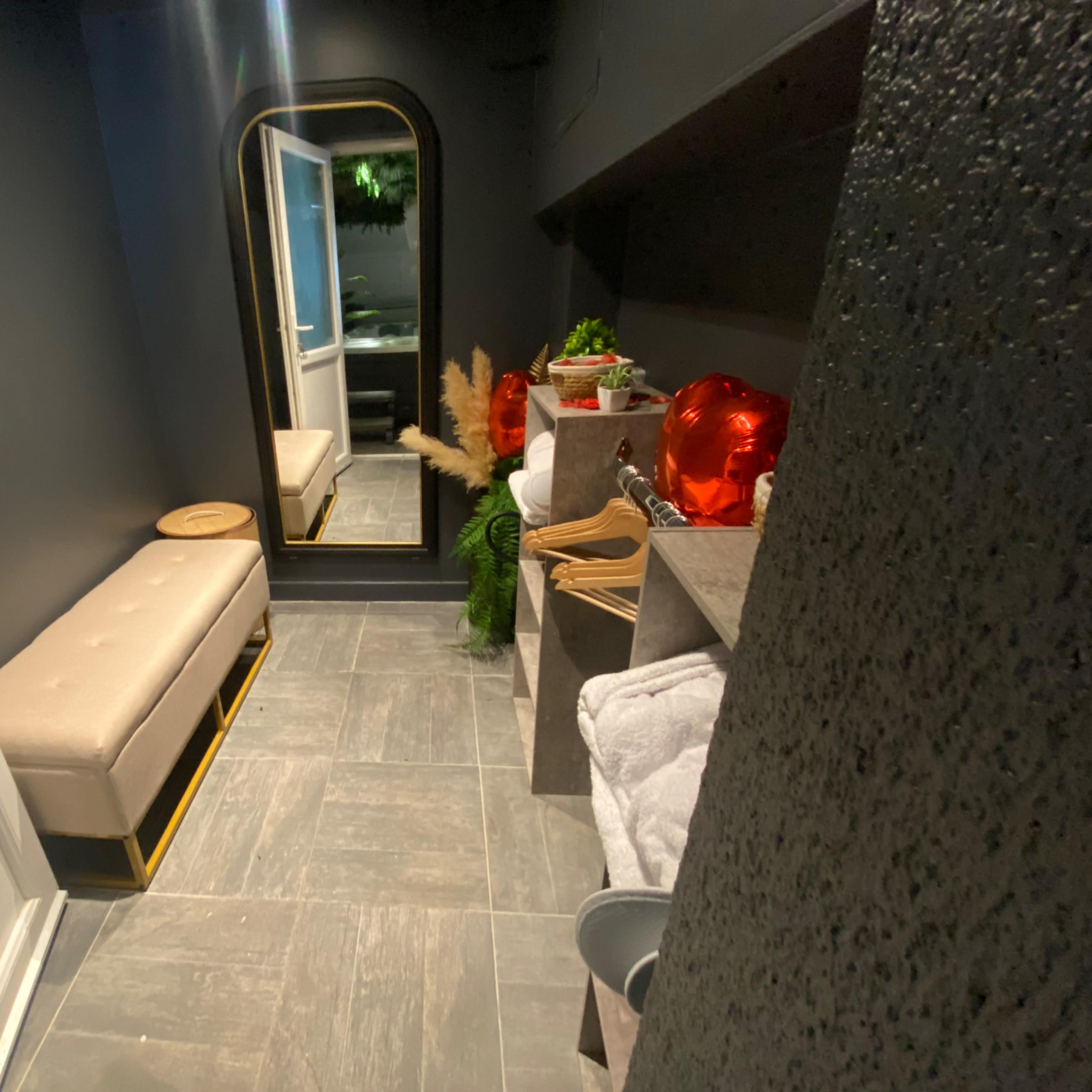 A narrow hallway with a bench, a large mirror, and shelves containing plants and towels against dark walls.