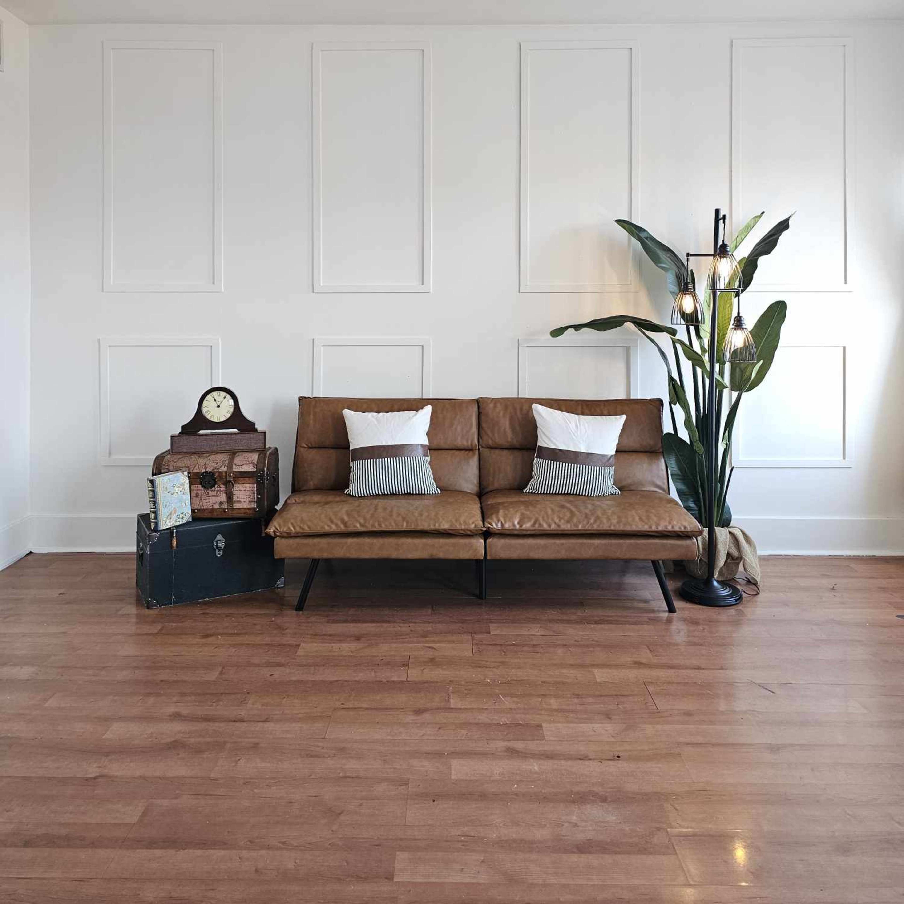 A mid-century leather sofa with white and striped pillows is positioned in a bright room with a large plant, a vintage clock, and wooden flooring.