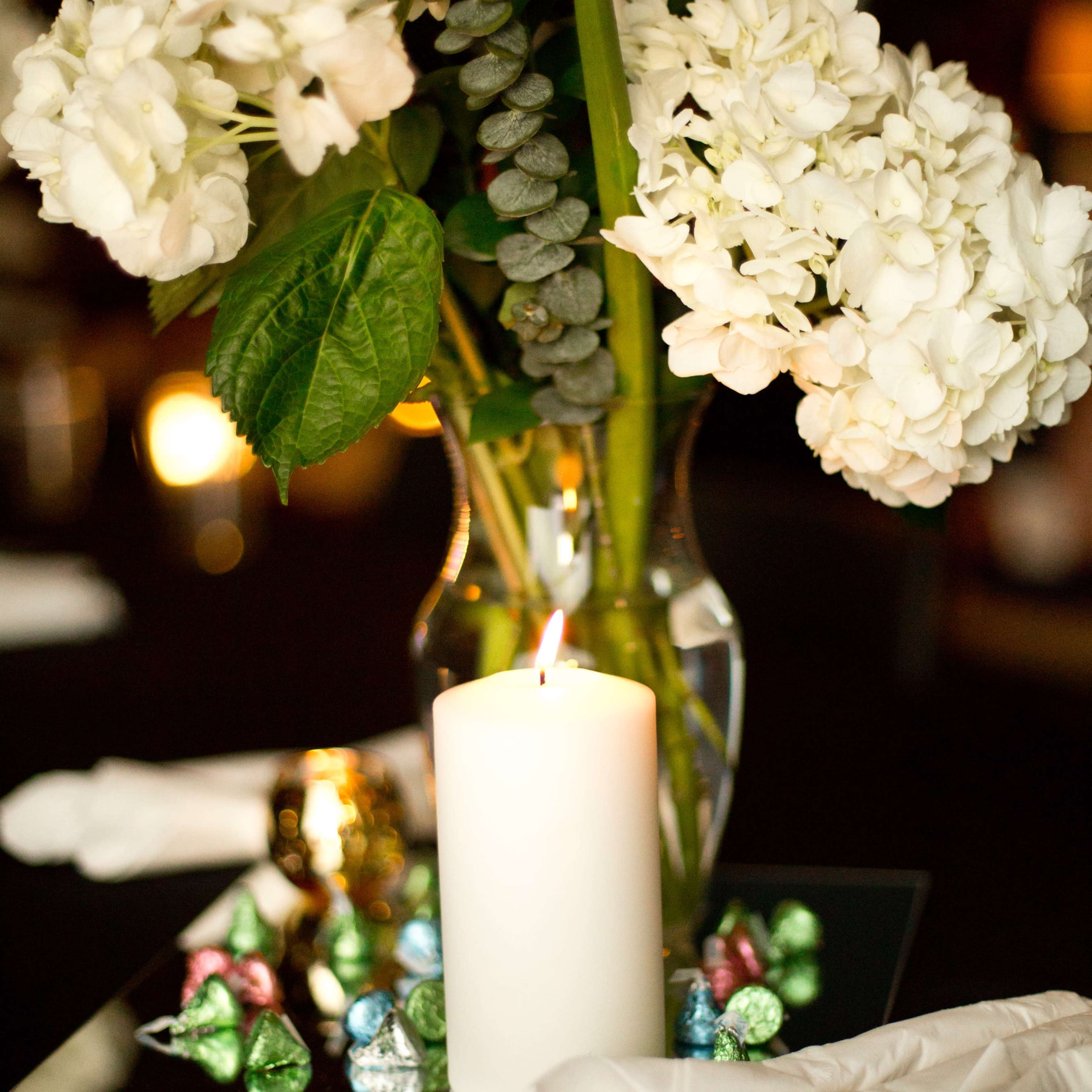 A vase of white flowers is placed on a black tablecloth, with a lit candle and colorful chocolates arranged around a mirrored base.